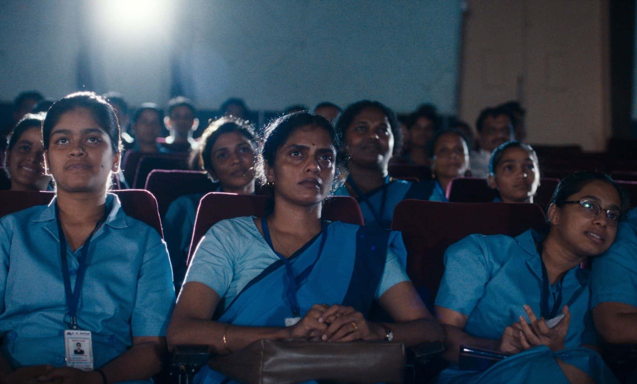 Rows of South Asian women dressed in blue sit in a theater, with a light shining in the background.