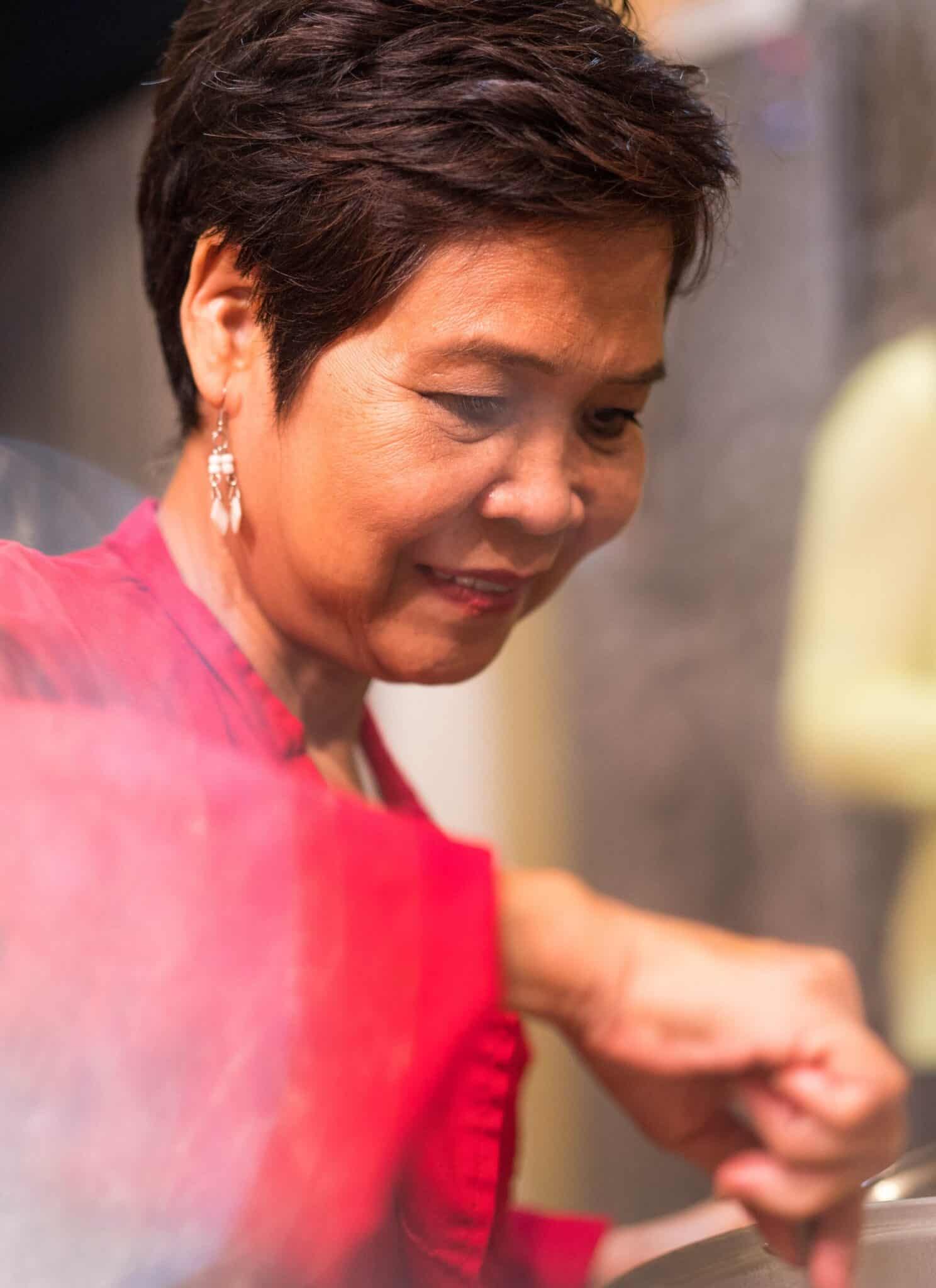An Asian woman with a pixie haircut, dressed in red, smiles while looking at a steaming pot.