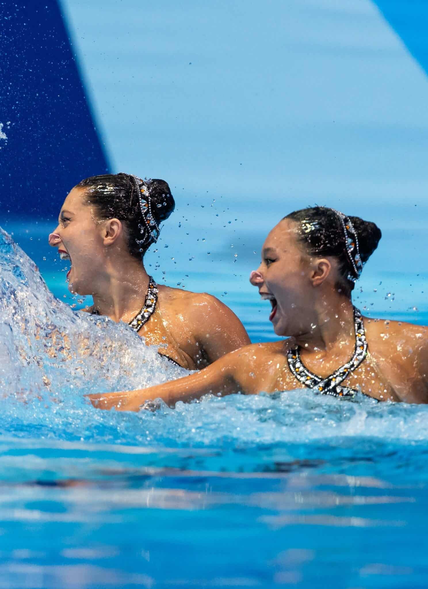 Artistic swimmers Ruby Remati and Megumi Field float in a pool, looking to the left, with a big splash of water in front of them.