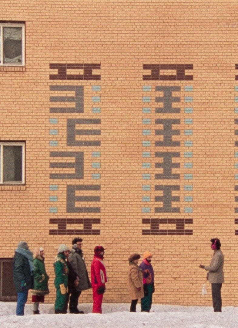 A row of people stand facing one man, against a light brown brick building.