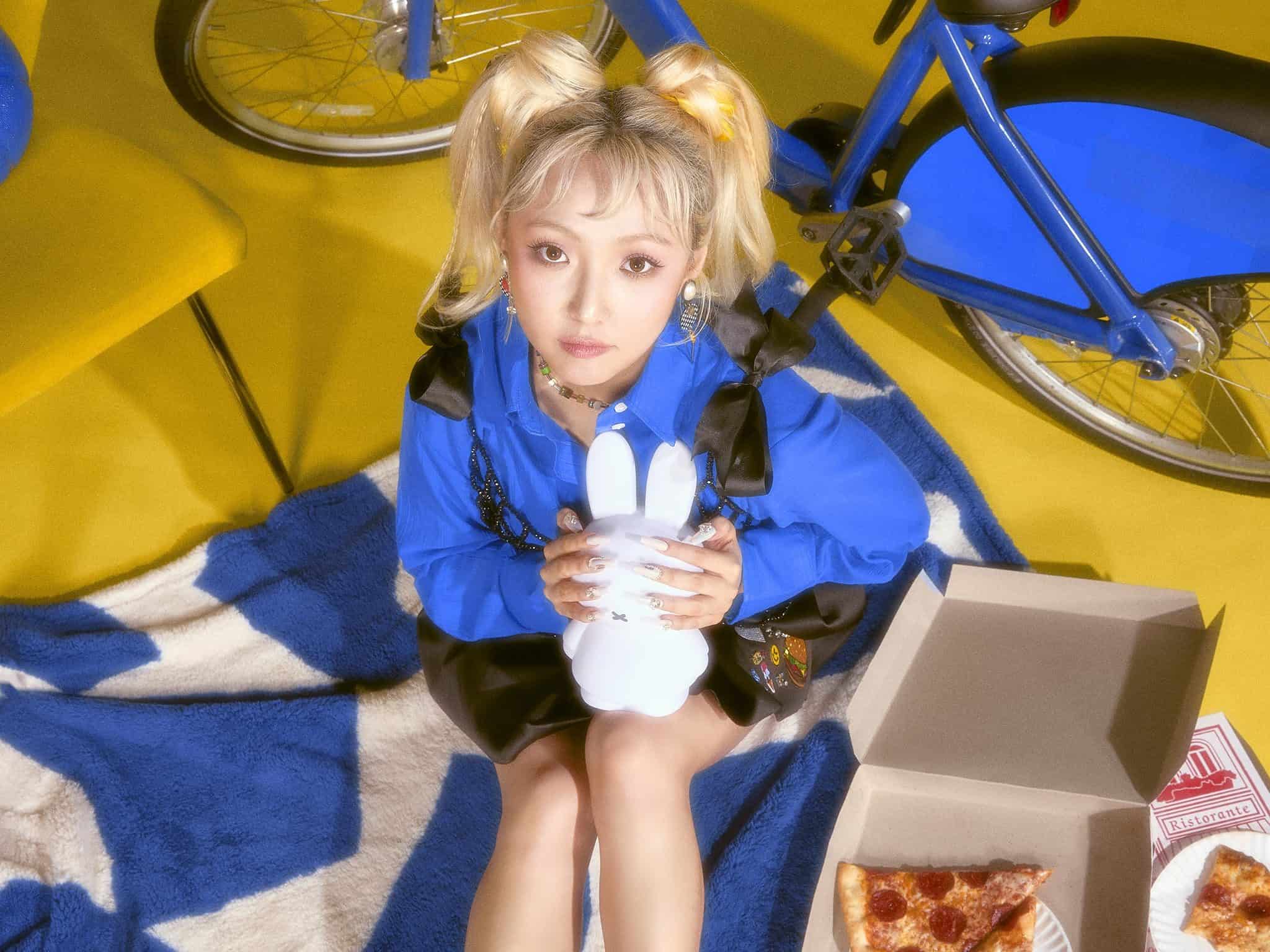 Singer Min, with blonde hair in two pigtails, dressed in blue, holds a white bunny and sits on a blue blanket, with a box of pizza next to her.