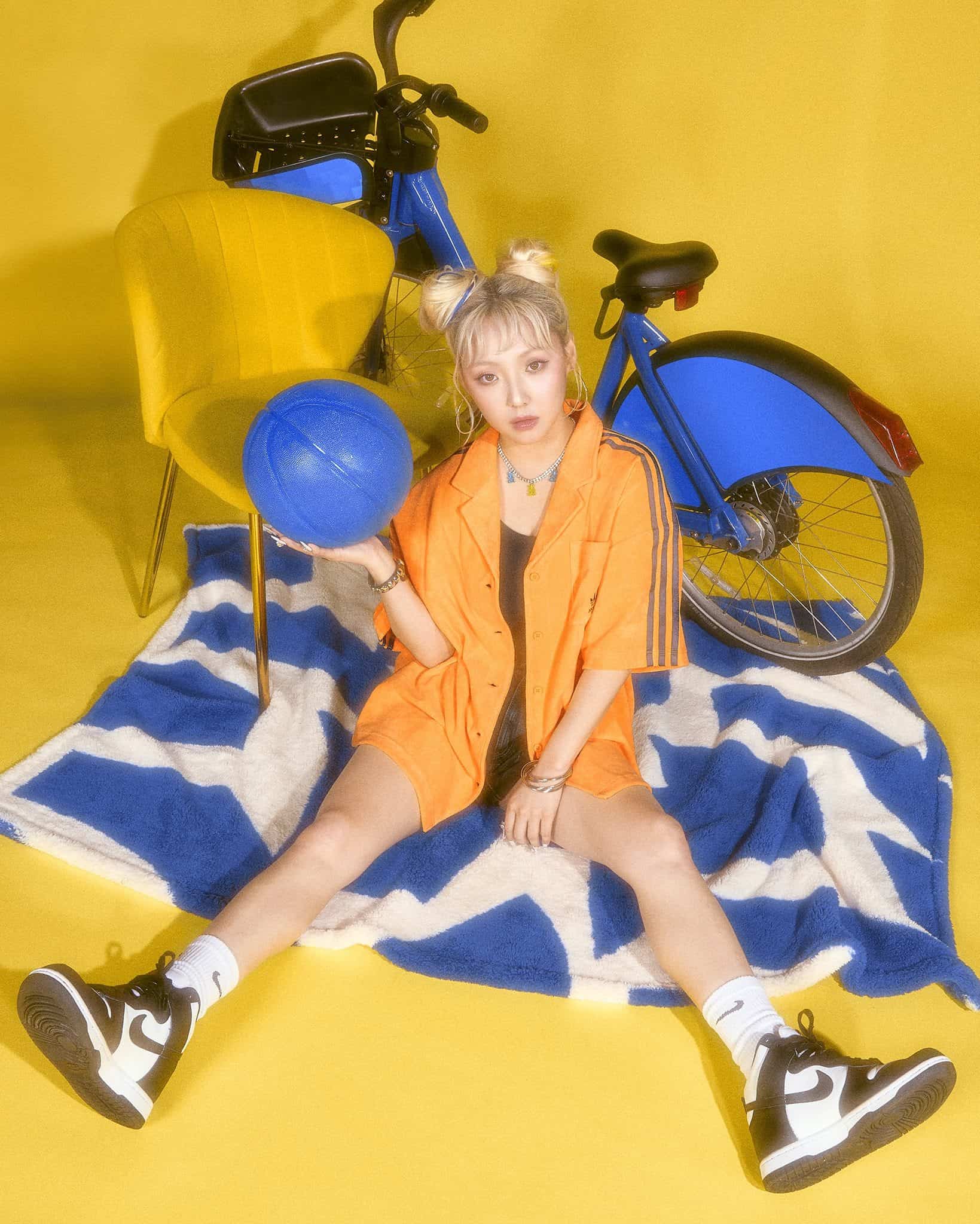 Singer Min, in an orange shirt and shorts, with blonde hair in two buns, sits on a blue blanket holding a blue ball and blue bike behind her against a yellow background.