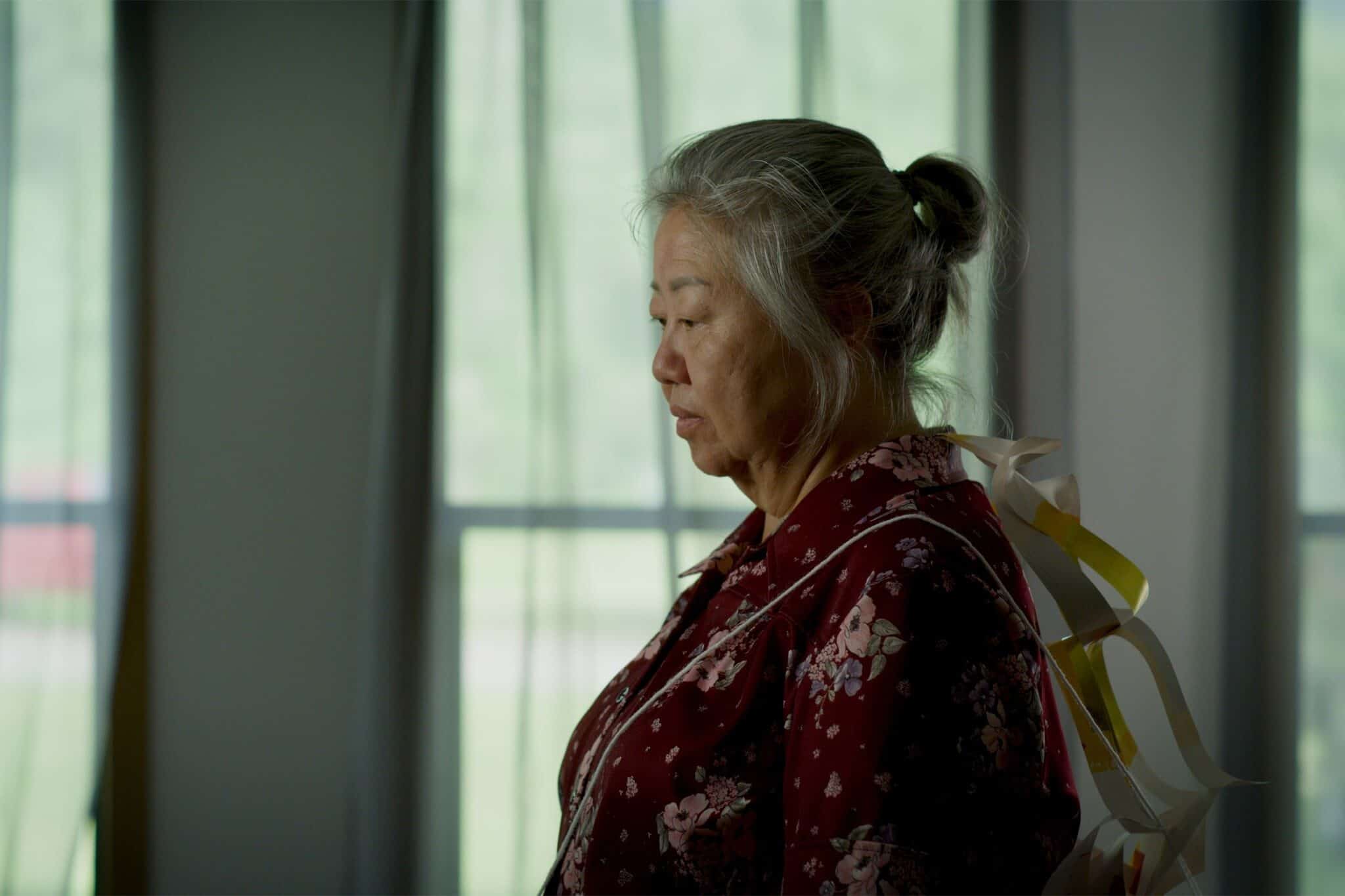 An older Asian woman, with gray hair in a ponytail, and a dark pink floral top, against a windowed wall in the background.