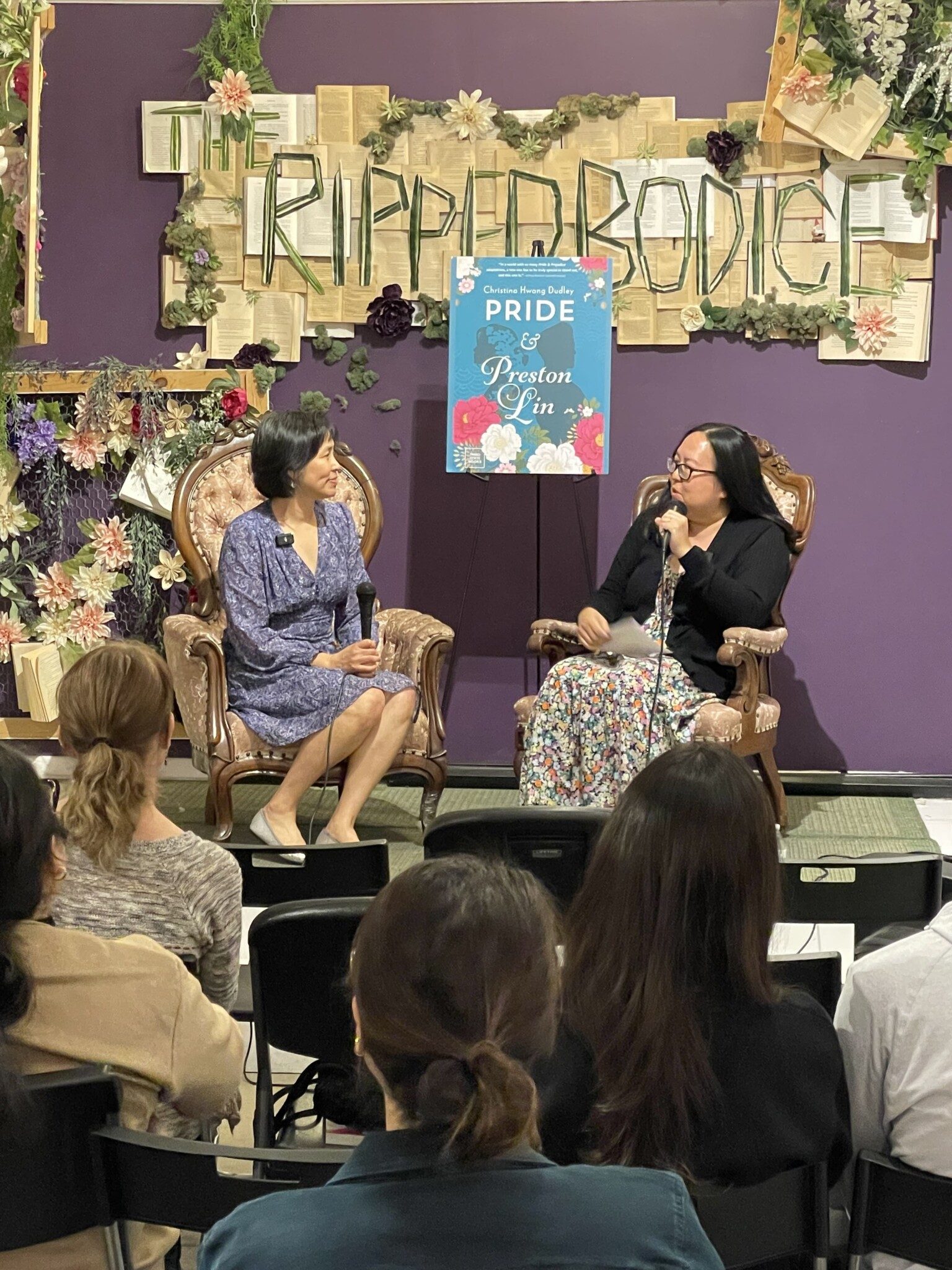 Two Asian women sit on chairs on a stage in front of an audience, with microphones, against a purple wall with a sign the reads "The Ripped Bodice."
