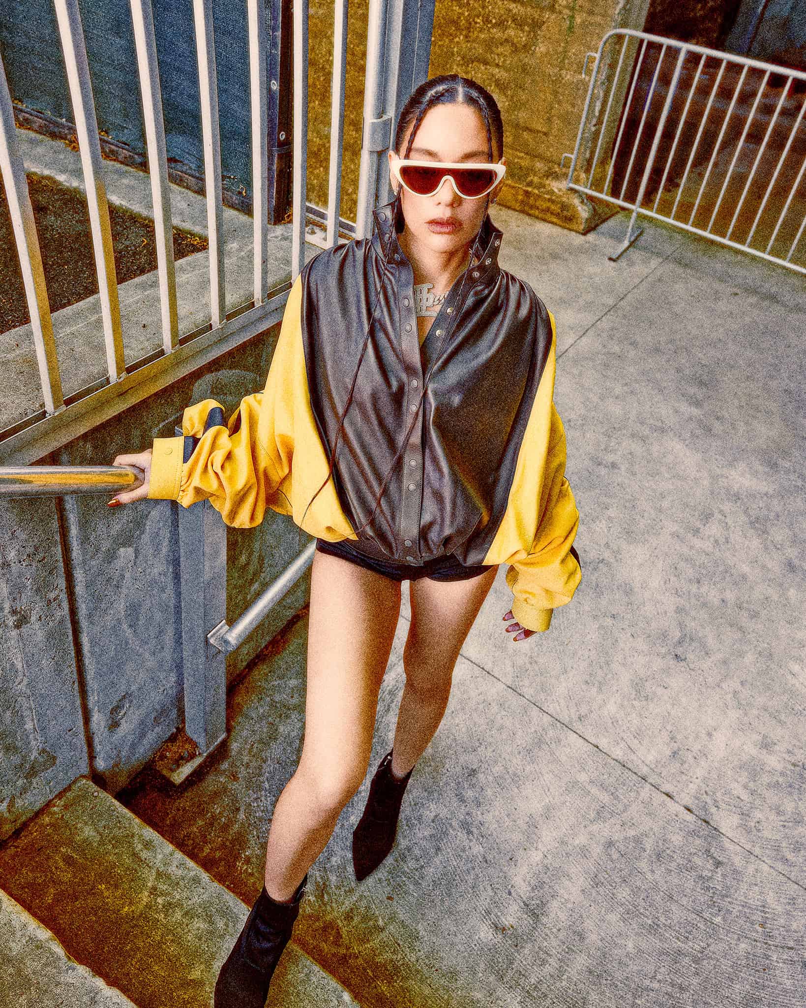 Japanese rapper AWICH, in a black and yellow jacket, shorts and white sunglasses, stands with gray railings in the background.