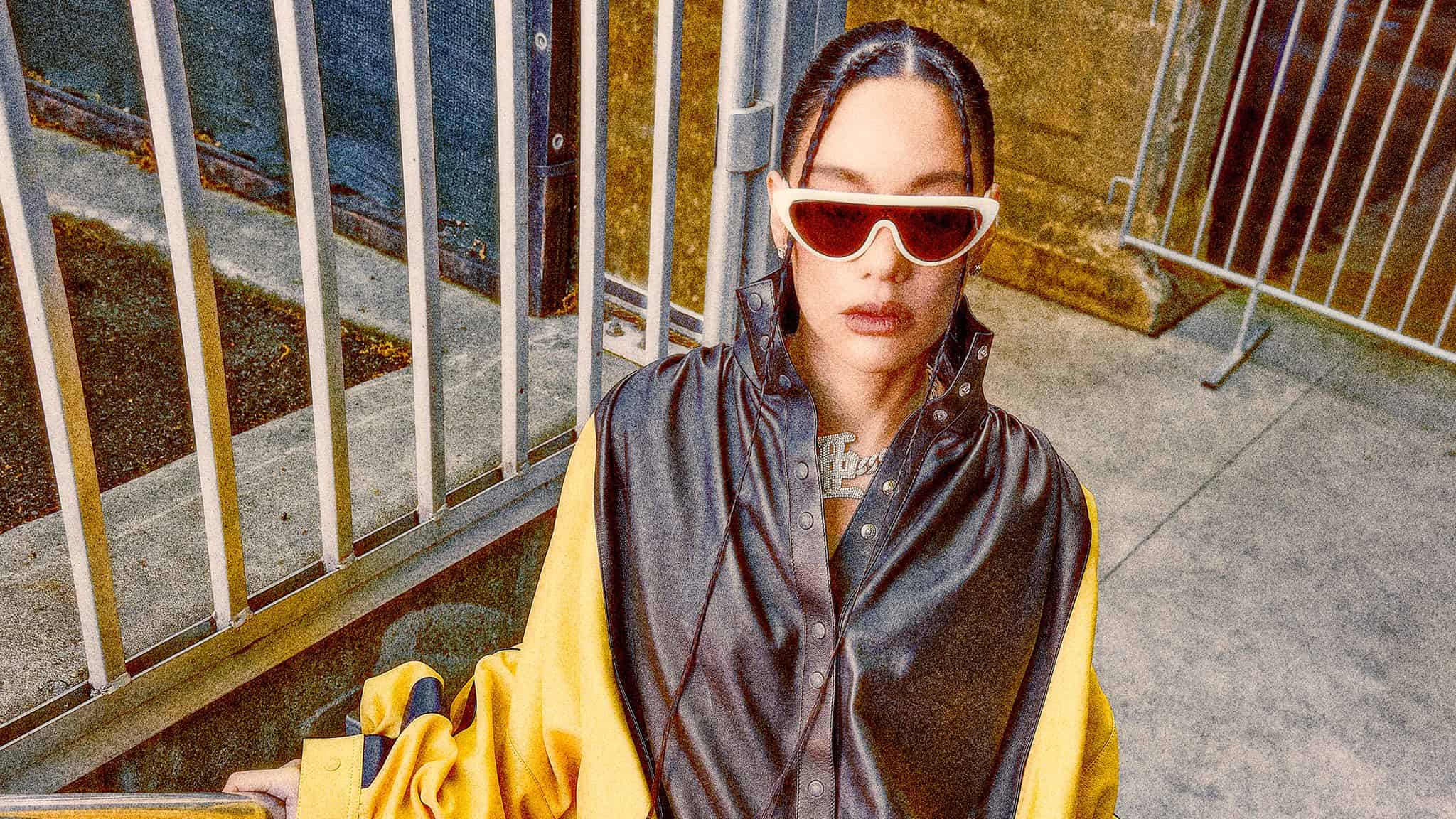 Japanese rapper AWICH, in a black and yellow jacket, shorts and white sunglasses, stands with gray railings in the background.