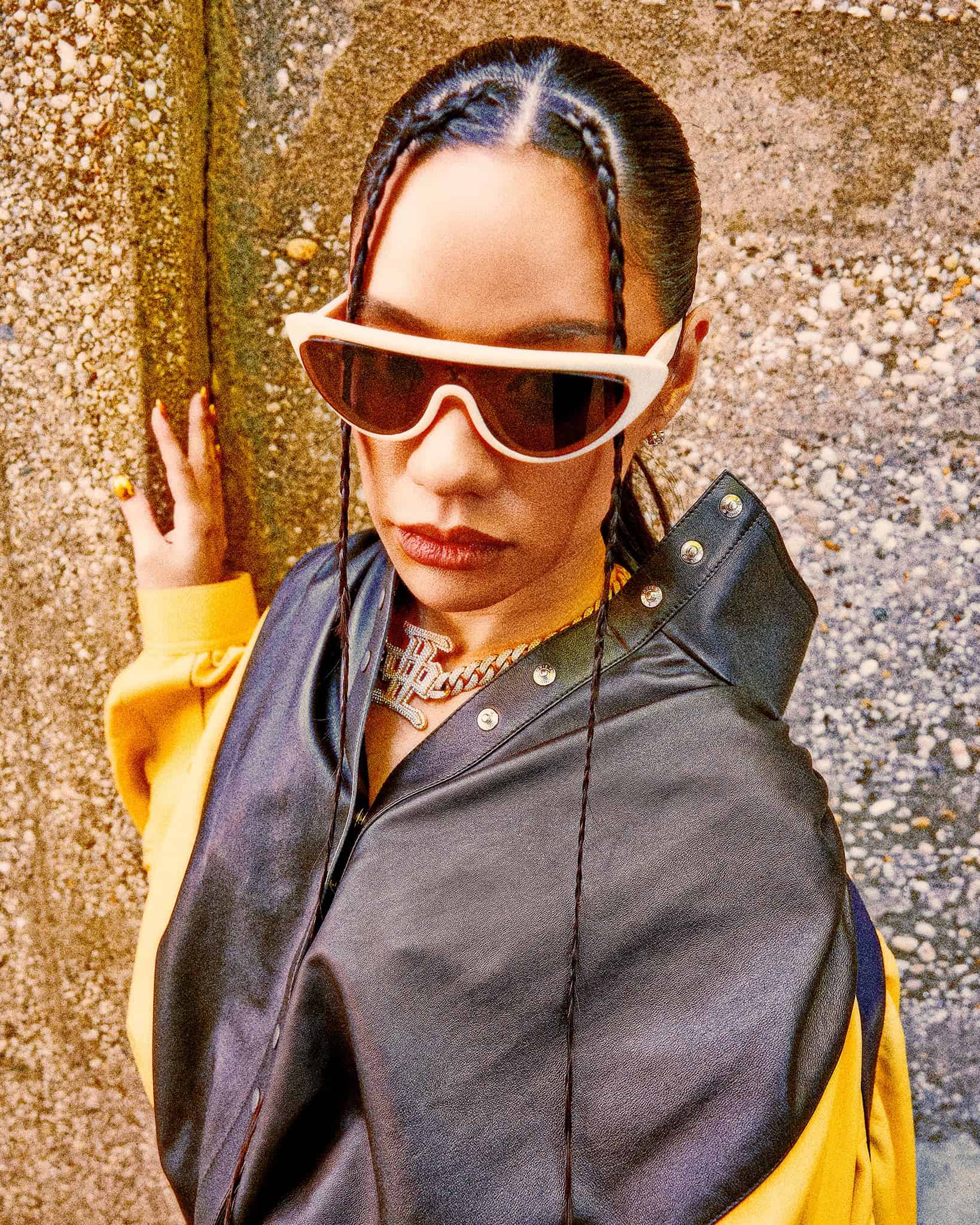 Japanese rapper AWICH, in a black and yellow jacket and white sunglasses, with her hand on a gray cement wall.