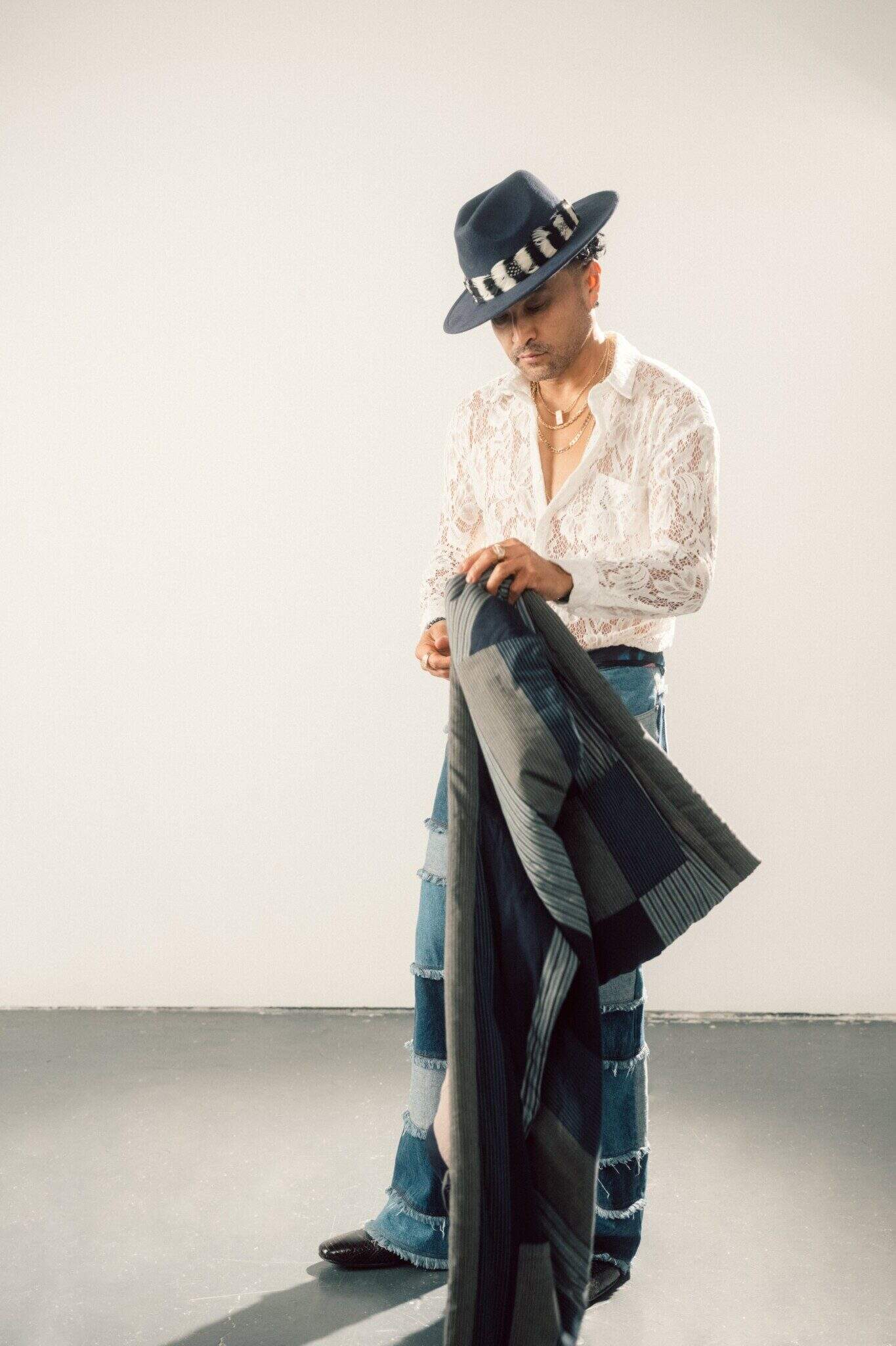 Rapper Lyrics Born, in a dark gray hat, white lace shirt and patchwork jeans, holds a long jacket against a white background.