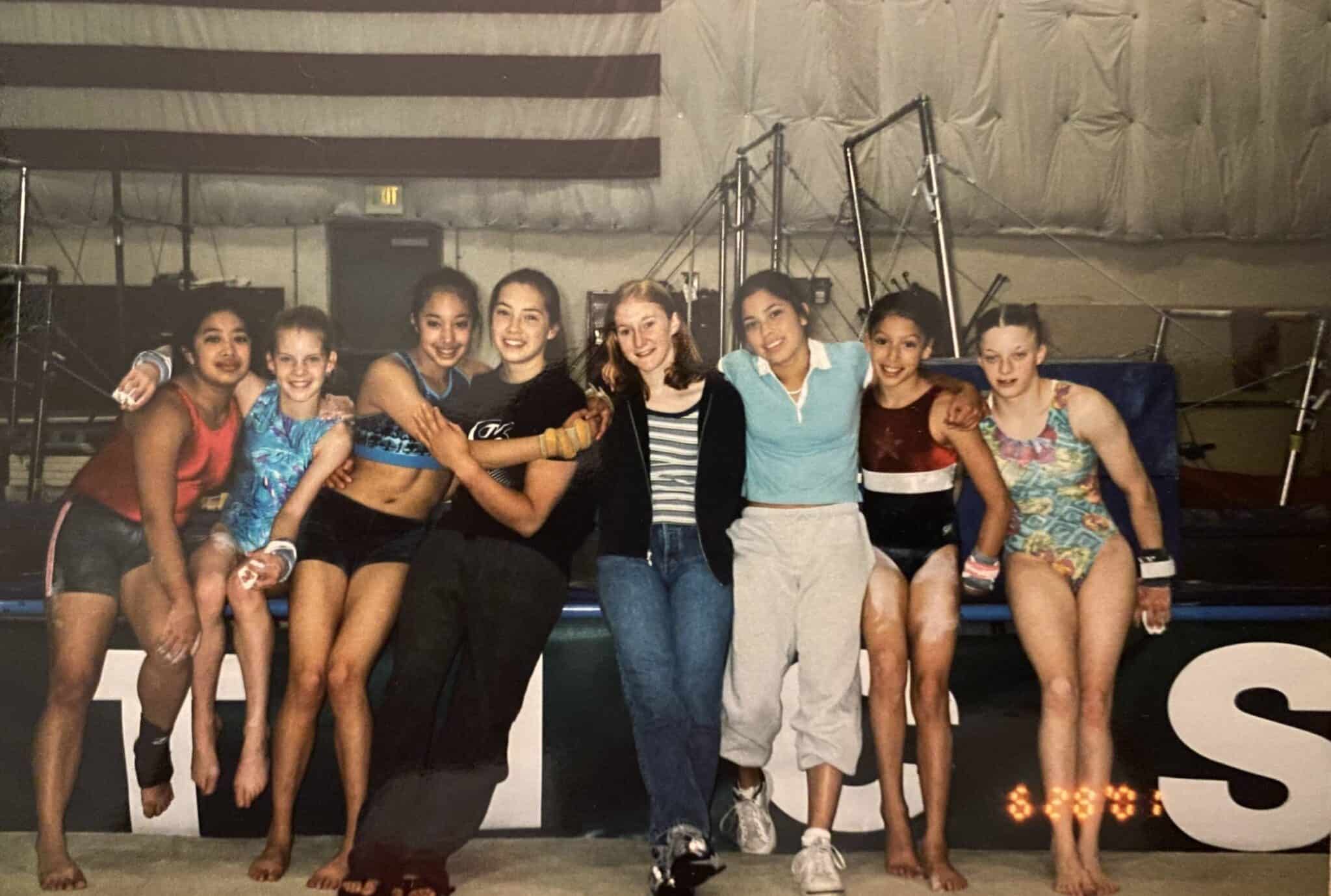 A row of young women stand together with gymnastics uneven bars in the background.