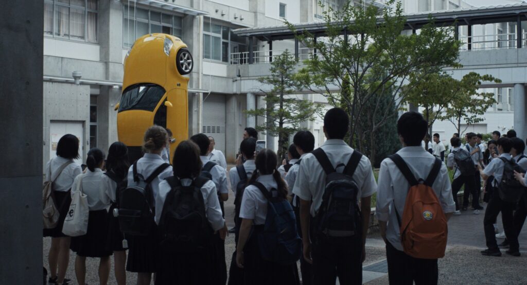 Still from "Happyend." A group of students stand in a crowd, staring at a yellow car.