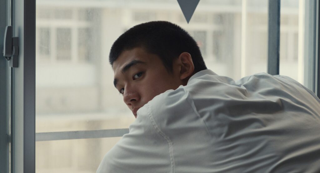 Still from "Happyend." A student in a white shirt leans near a window, he looks over his shoulder.