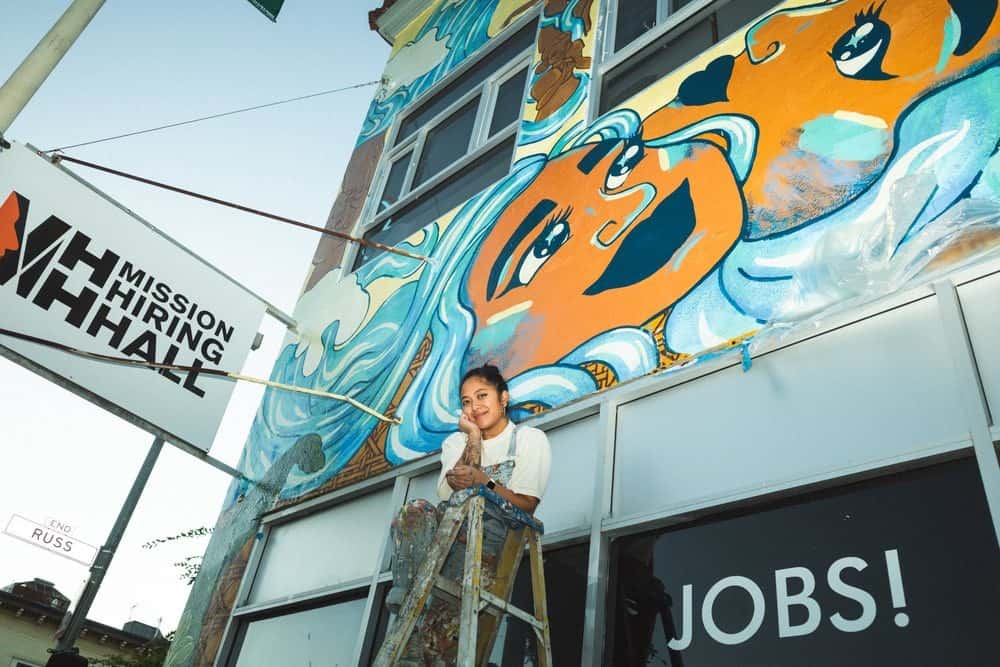 A woman smiling for the camera while standing on a ladder in front of a mural.