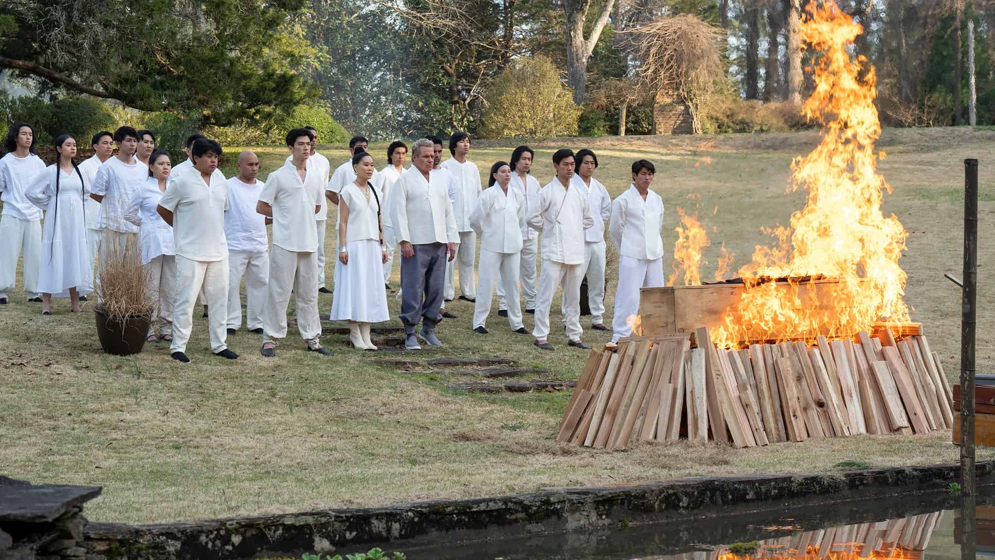 Still from "Cobra Kai" season six episode 11. A group of people stand staring at a burning coffin.