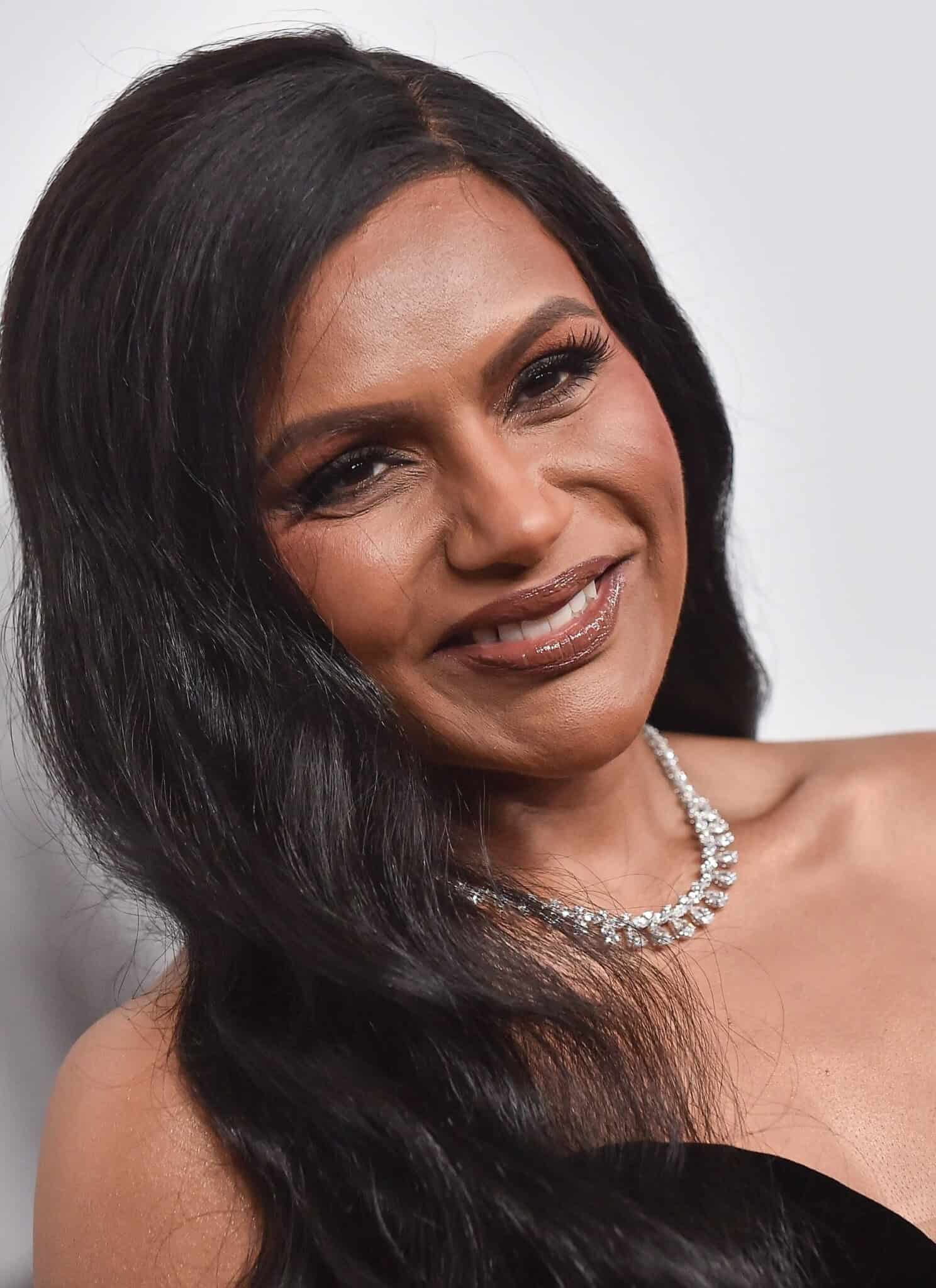 Actress Mindy Kaling, in a black dress, with her hair down and a diamond necklace, against a white background.