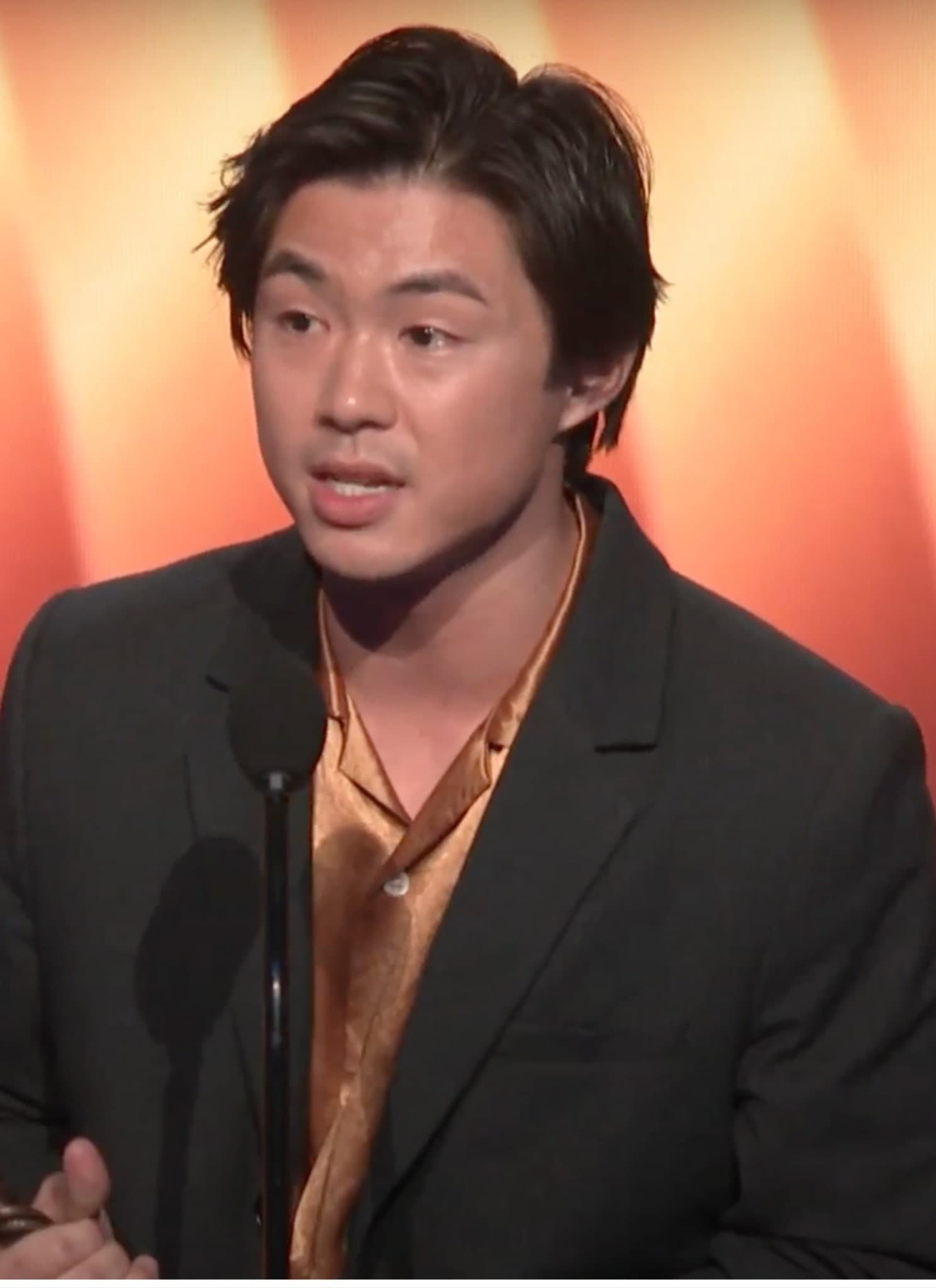 Sean Wang in black suit with orange shirt standing on stage at Film Independent Spirit Awards.