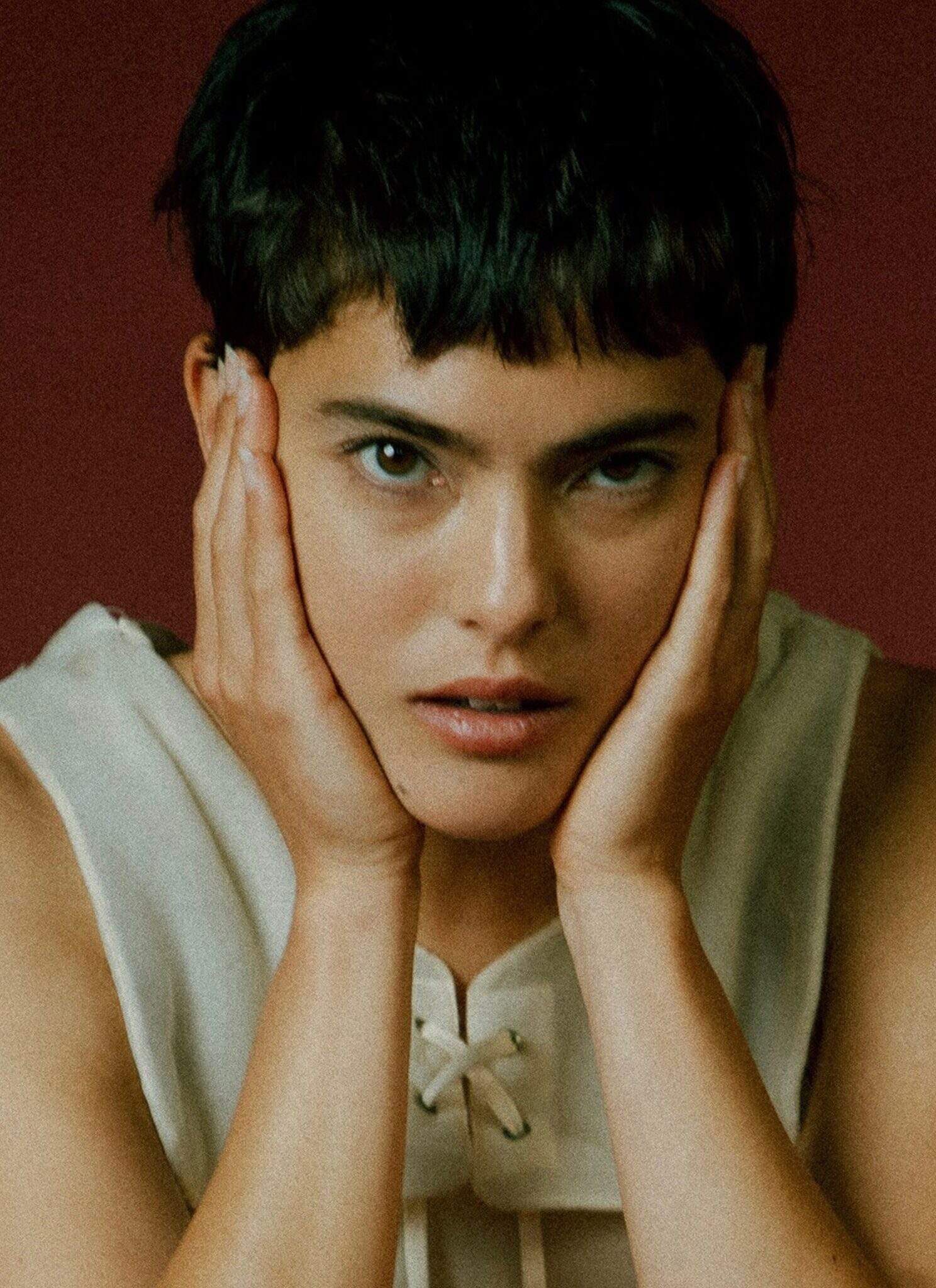 Miya Folick holding her face in her hands in front of a red background.