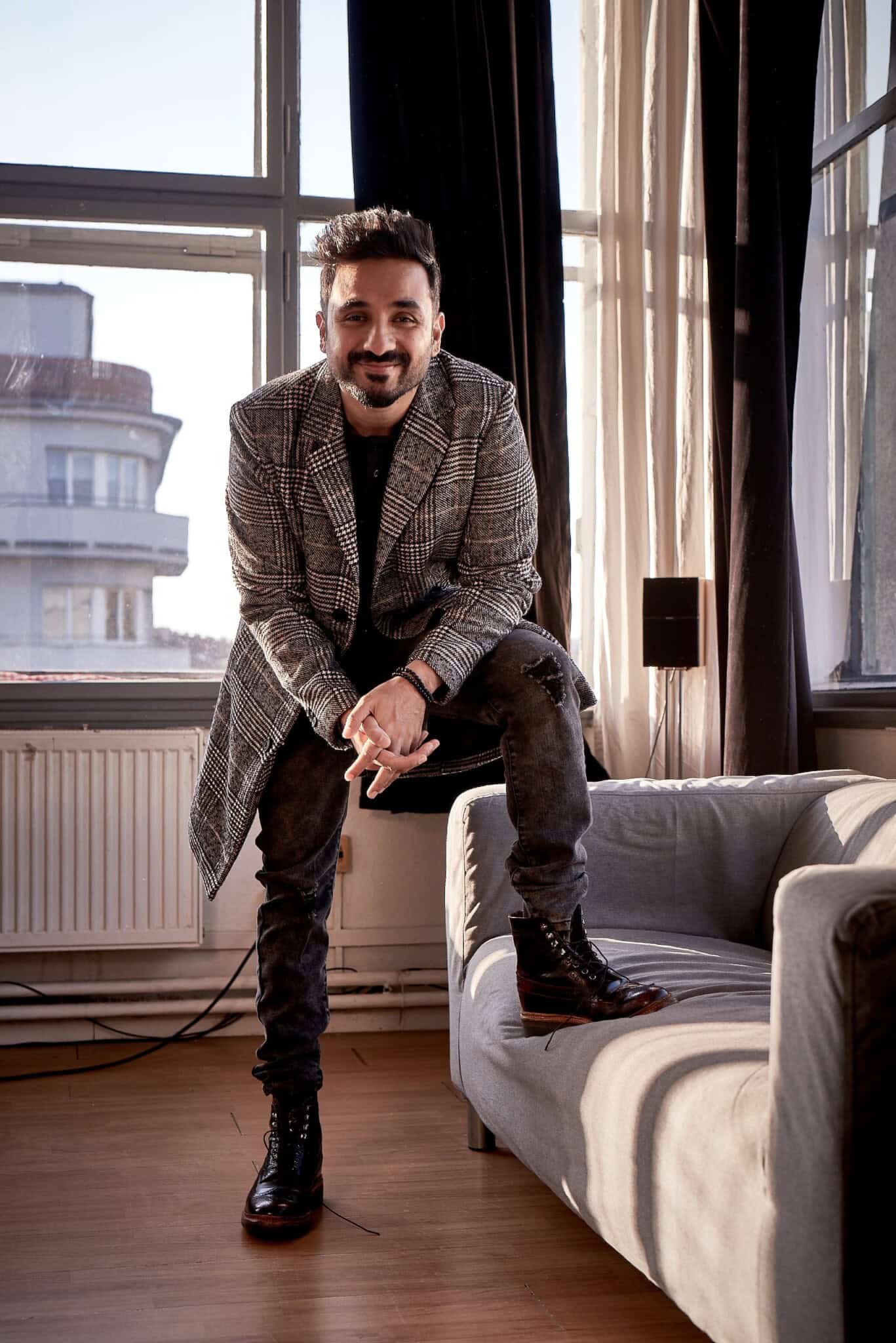 Vir Das standing in a room, one foot propped up on a couch.