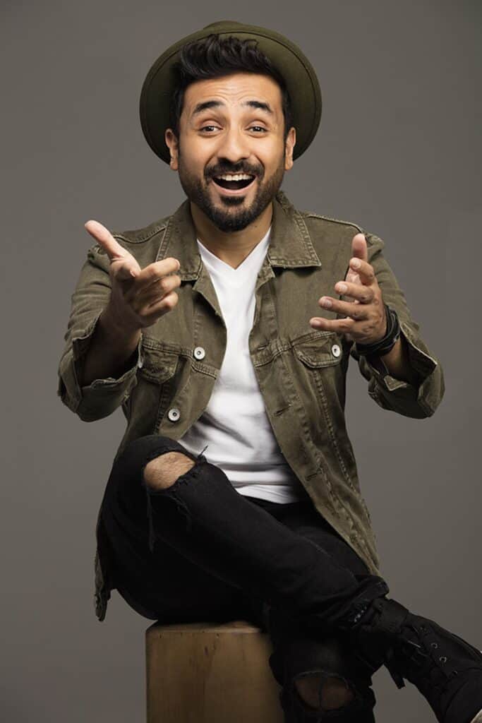 Vir Das sitting and smiling, his hands outstretched towards the camera.