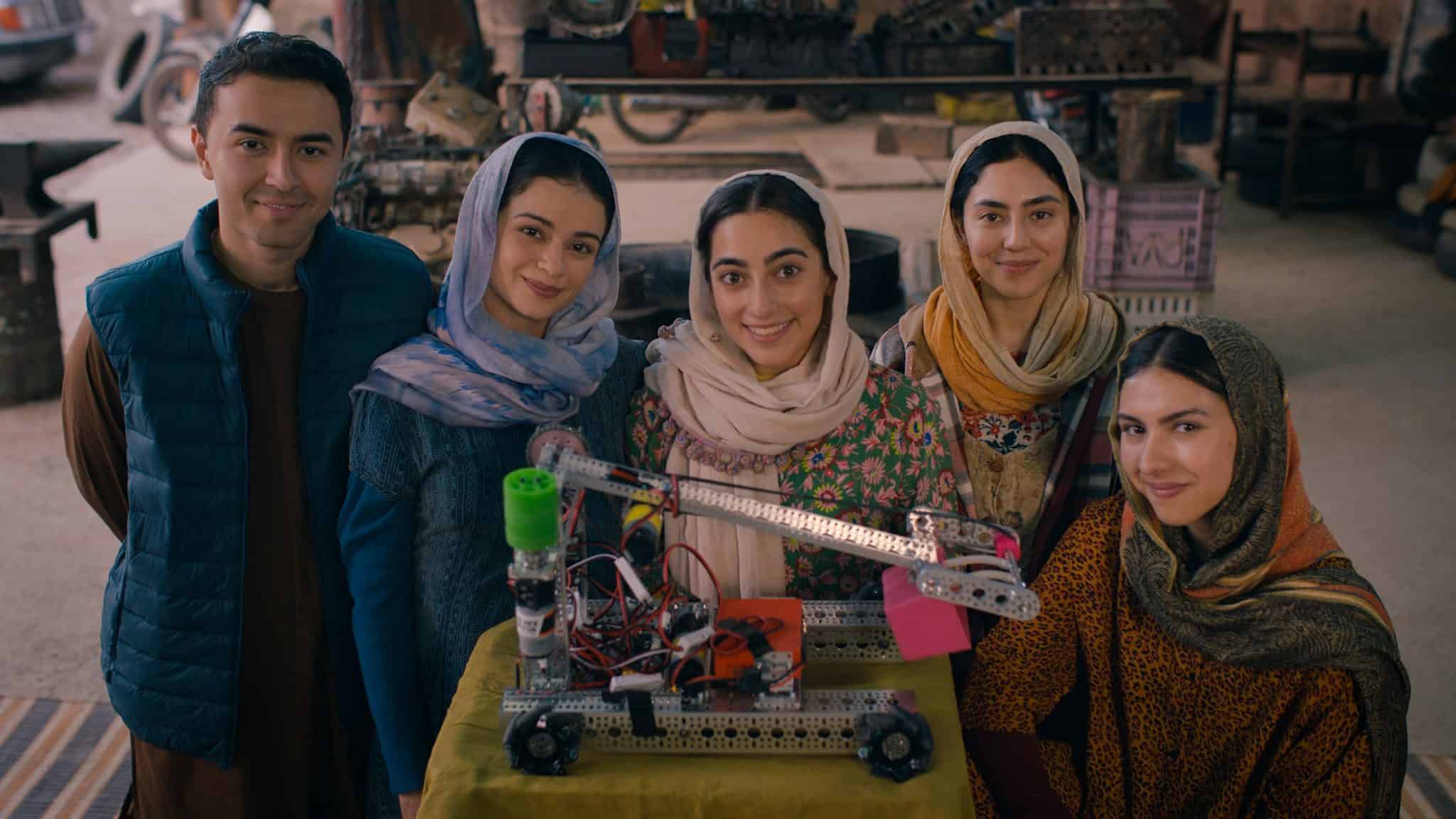 A group of women and a man smile at the camera, posing behind a small robot.