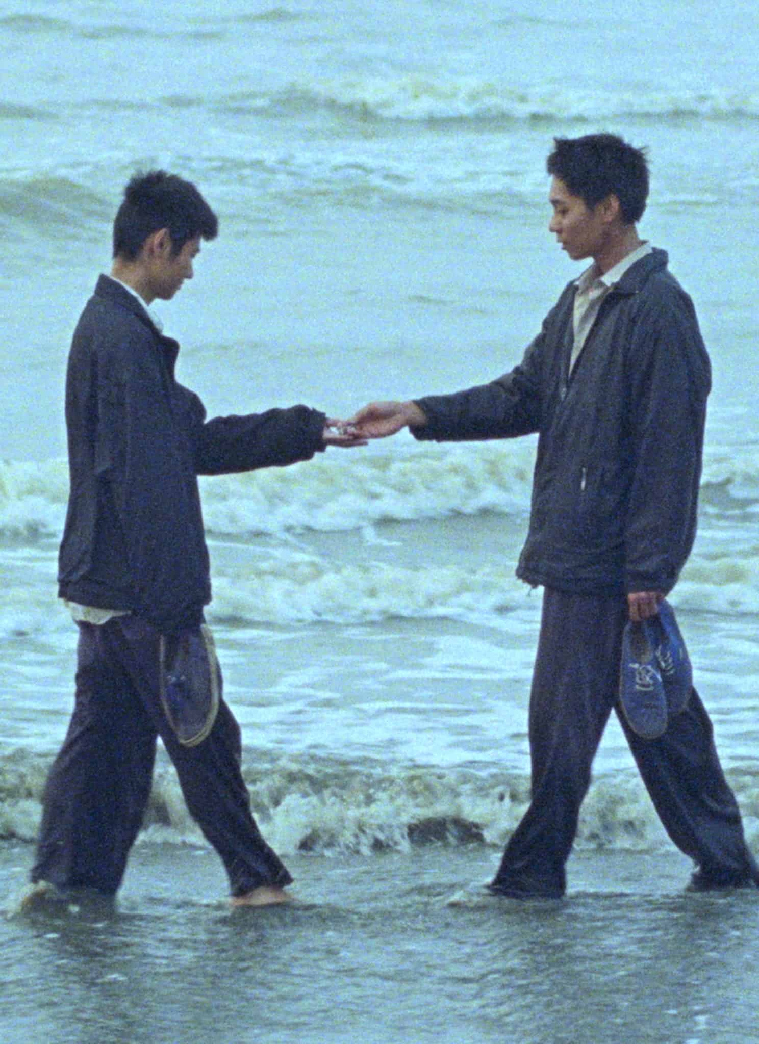 Still from "Viet and Nam"; two men stand on the beach, handing each other something.