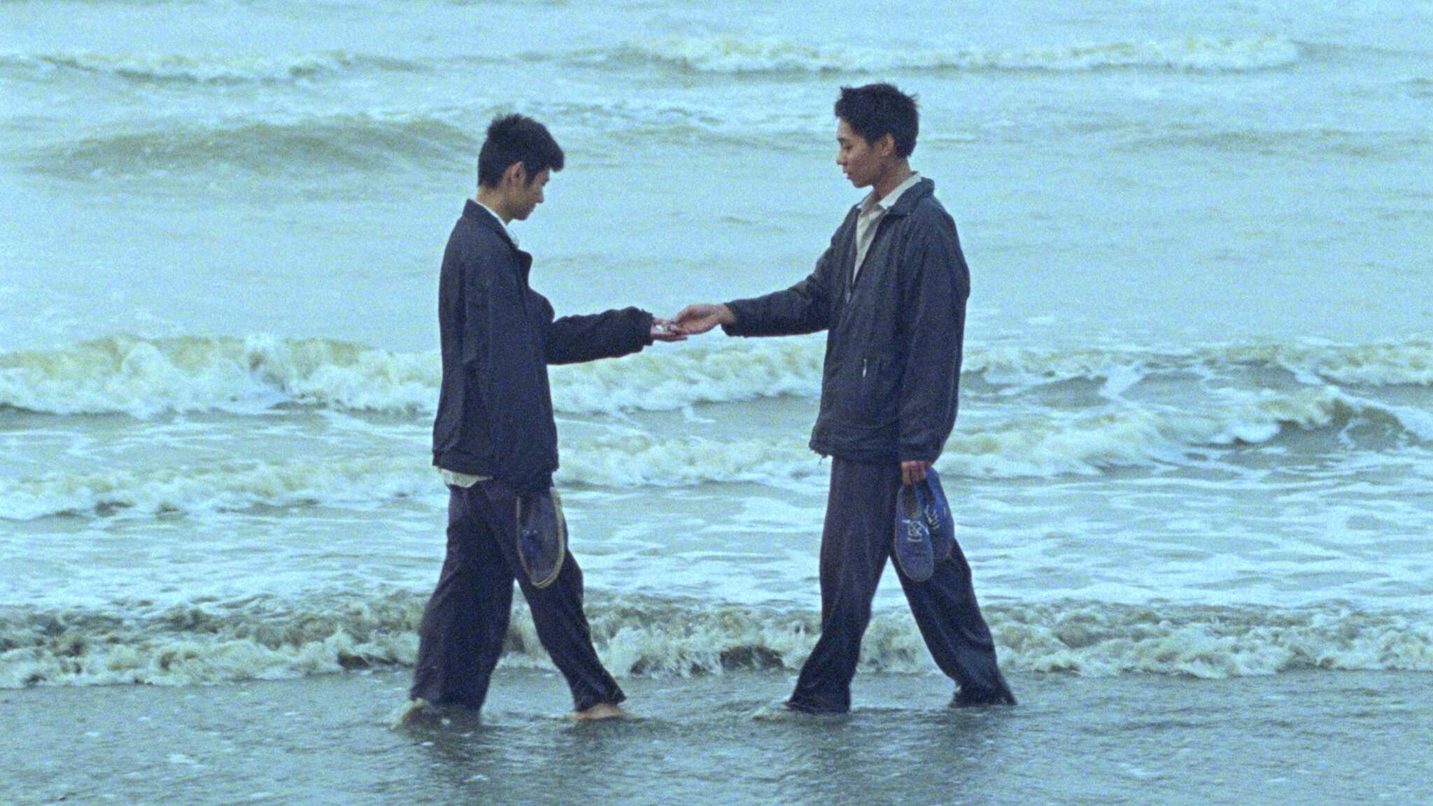 Still from "Viet and Nam"; two men stand on the beach, handing each other something.