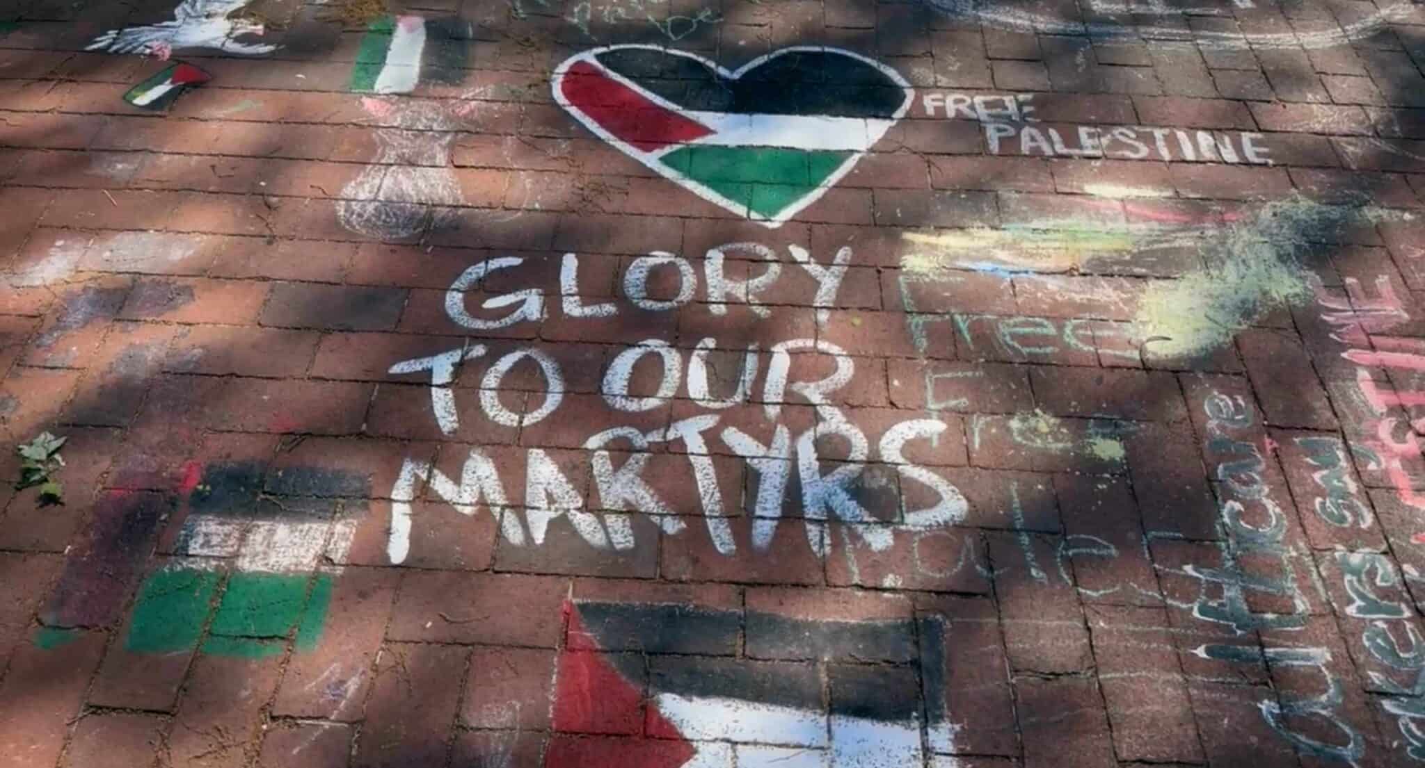 Chalk meesage on the floor reading "GLORY TO OUR MARTYRS" with the Palestine flag next to it.
