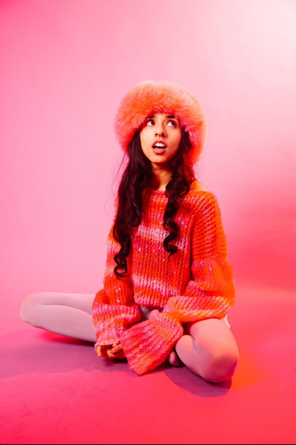 Misa Sattar in an orange and pink outfit sitting cross-legged in front of a pinkish, orange background.