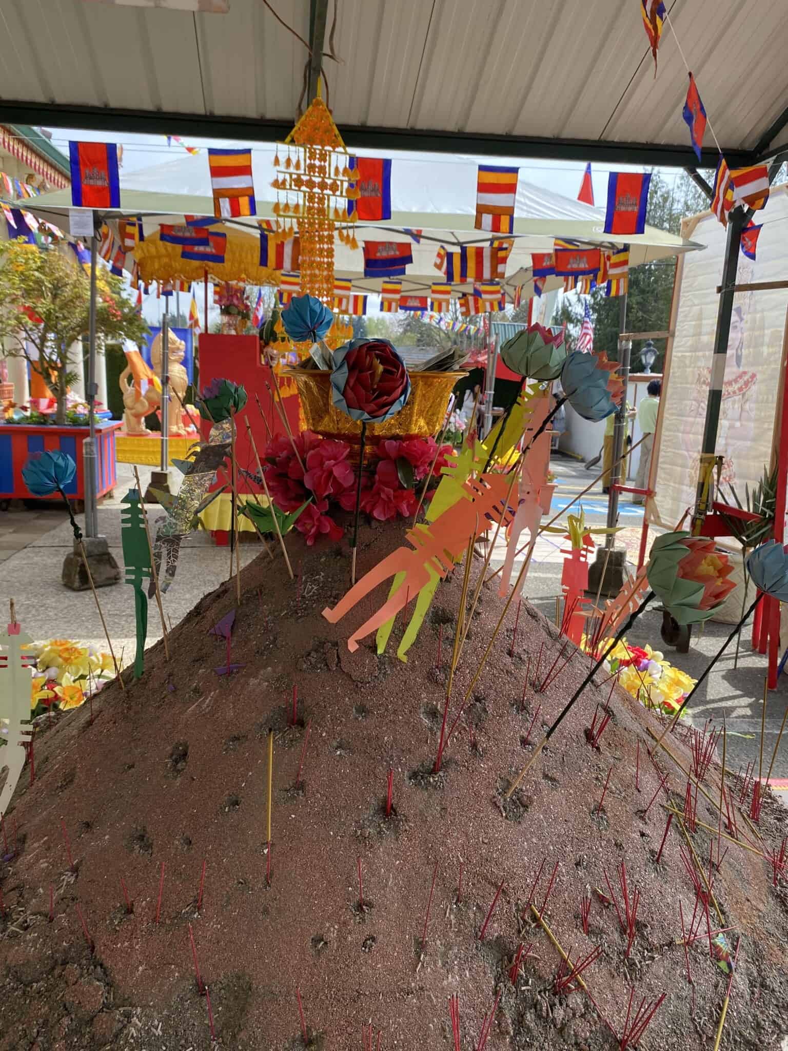 A mound of sand decorated with flags, flowers and incense, with small Cambodian flags and other colorful flags in the background.