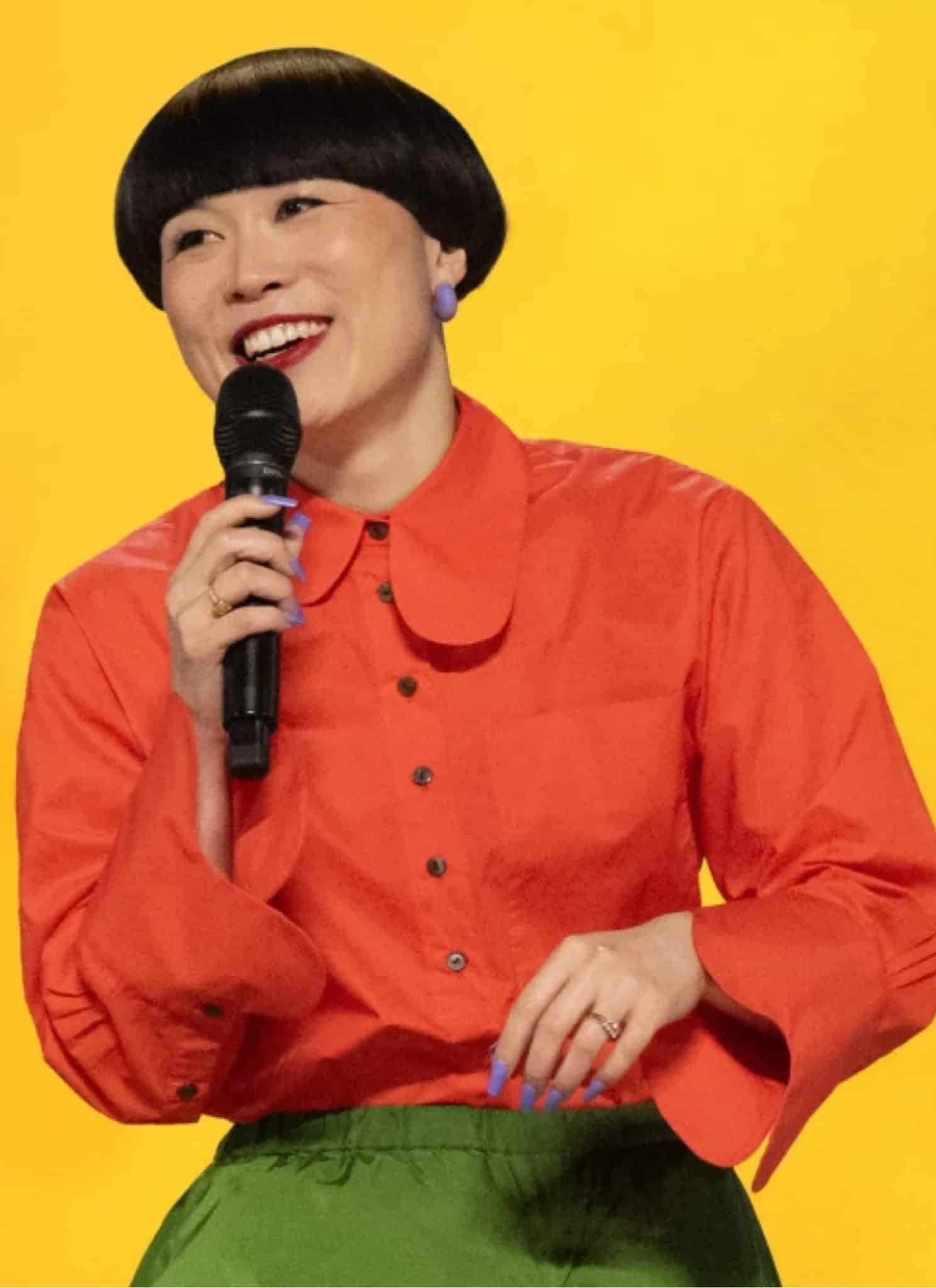 Atsuko in orange shirt with yellow background holding a mic.