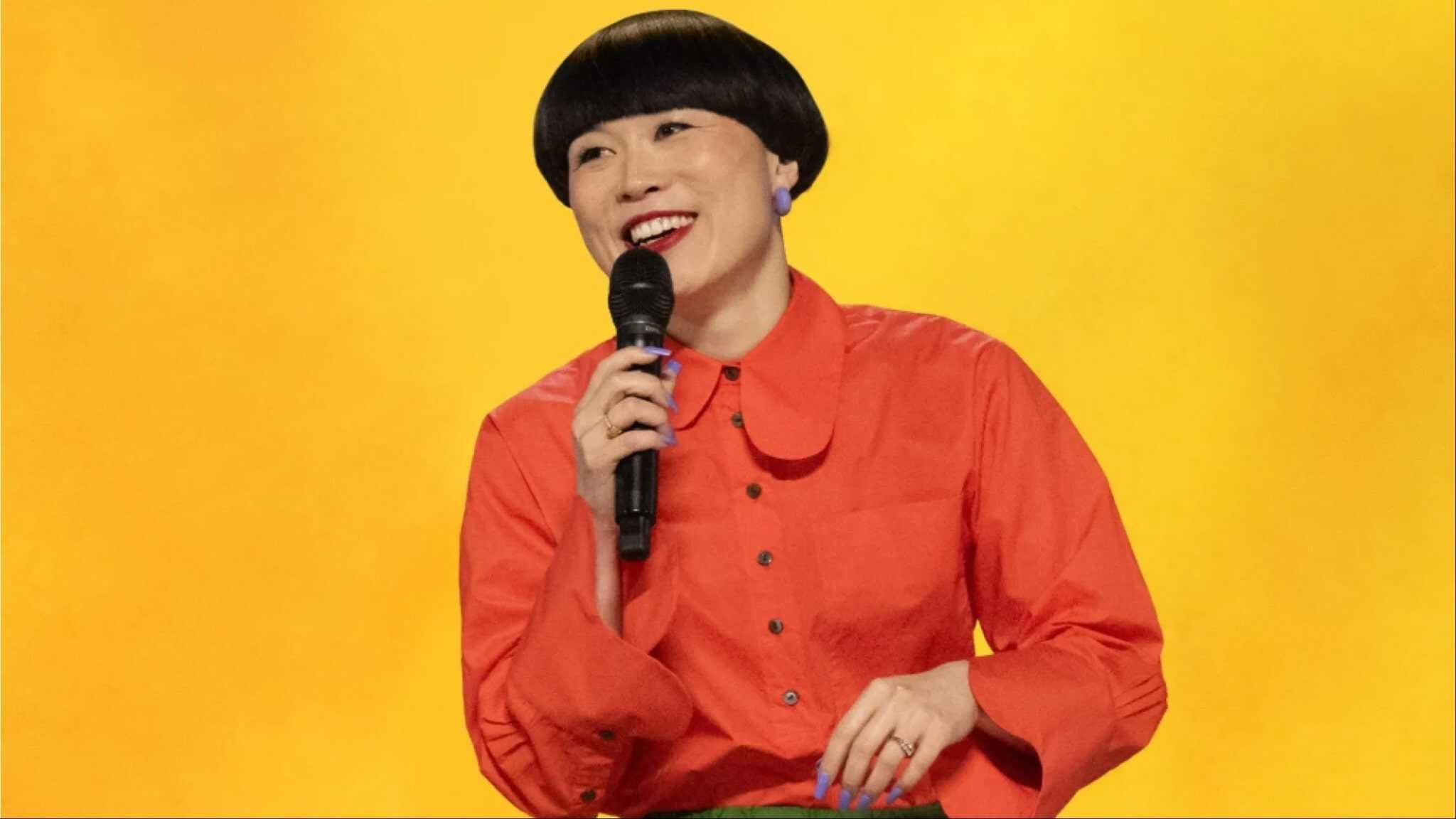 Atsuko in orange shirt with yellow background holding a mic.