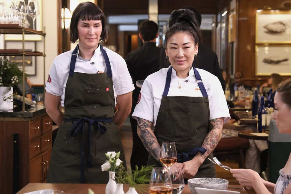 Chefs Katianna Hong and Sarah Levy stand together in white tops and black aprons at a table with bowls and glasses.