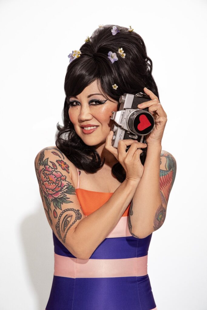Margaret Cho wearing a stripped dress and holding a film camera in her hands.
