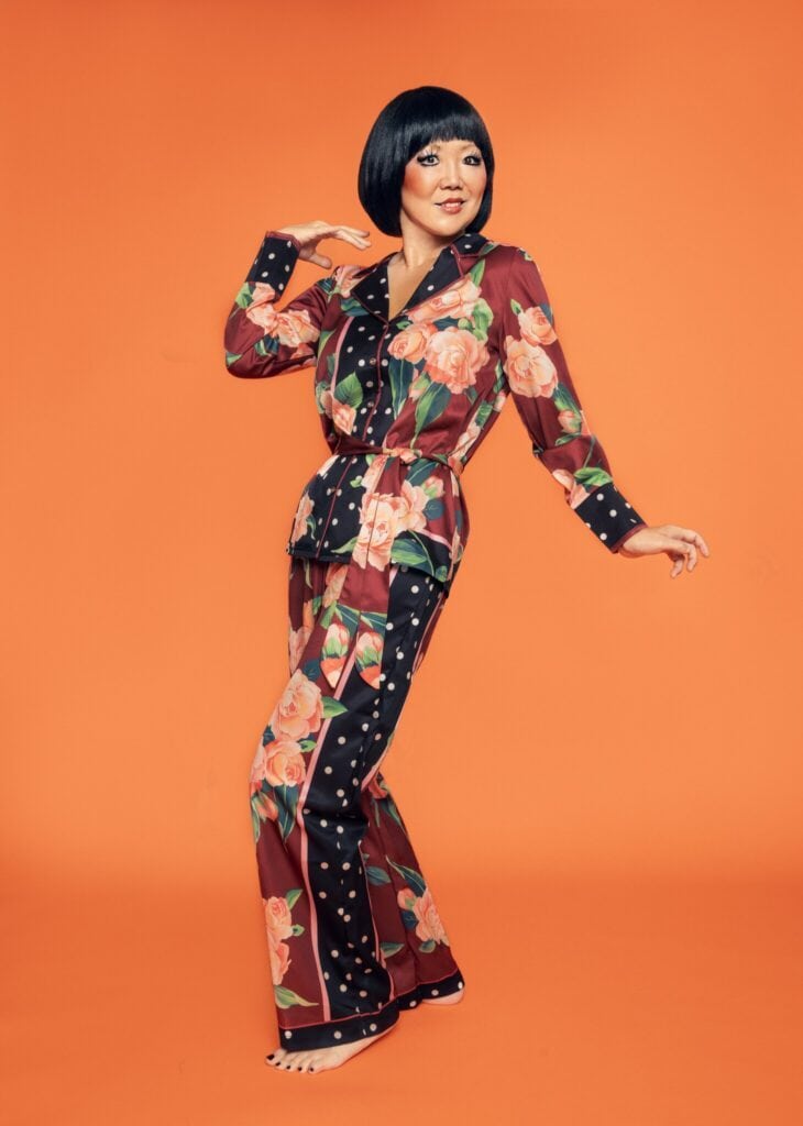 Margaret Cho in a patterned jumpsuit and a bob in front of an orange background.