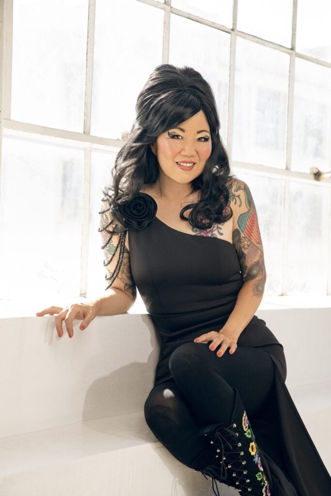 Margaret Cho in a black asymmetrical outfit sitting in front of a window.
