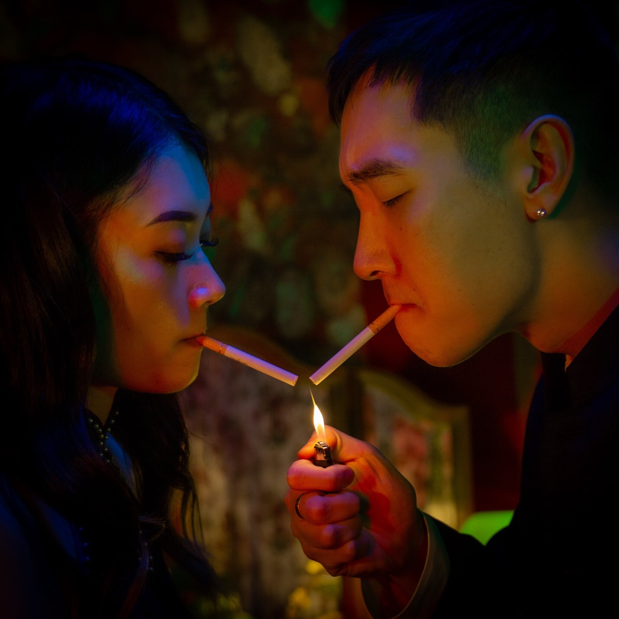 An Asian woman and man face each other with cigarettes in their mouths, with a lit lighter in between them.