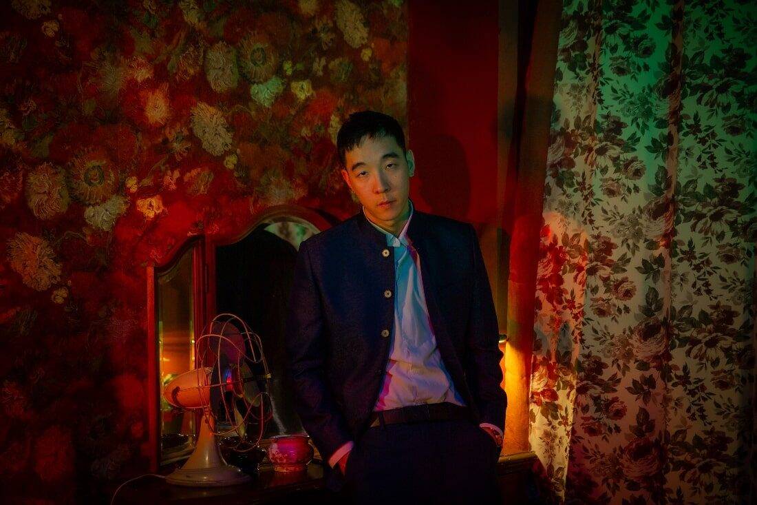 Chinese American rapper Chow Mane, dressed in a dark Chinese-style jacket in a "In the Mood for Love" styled room