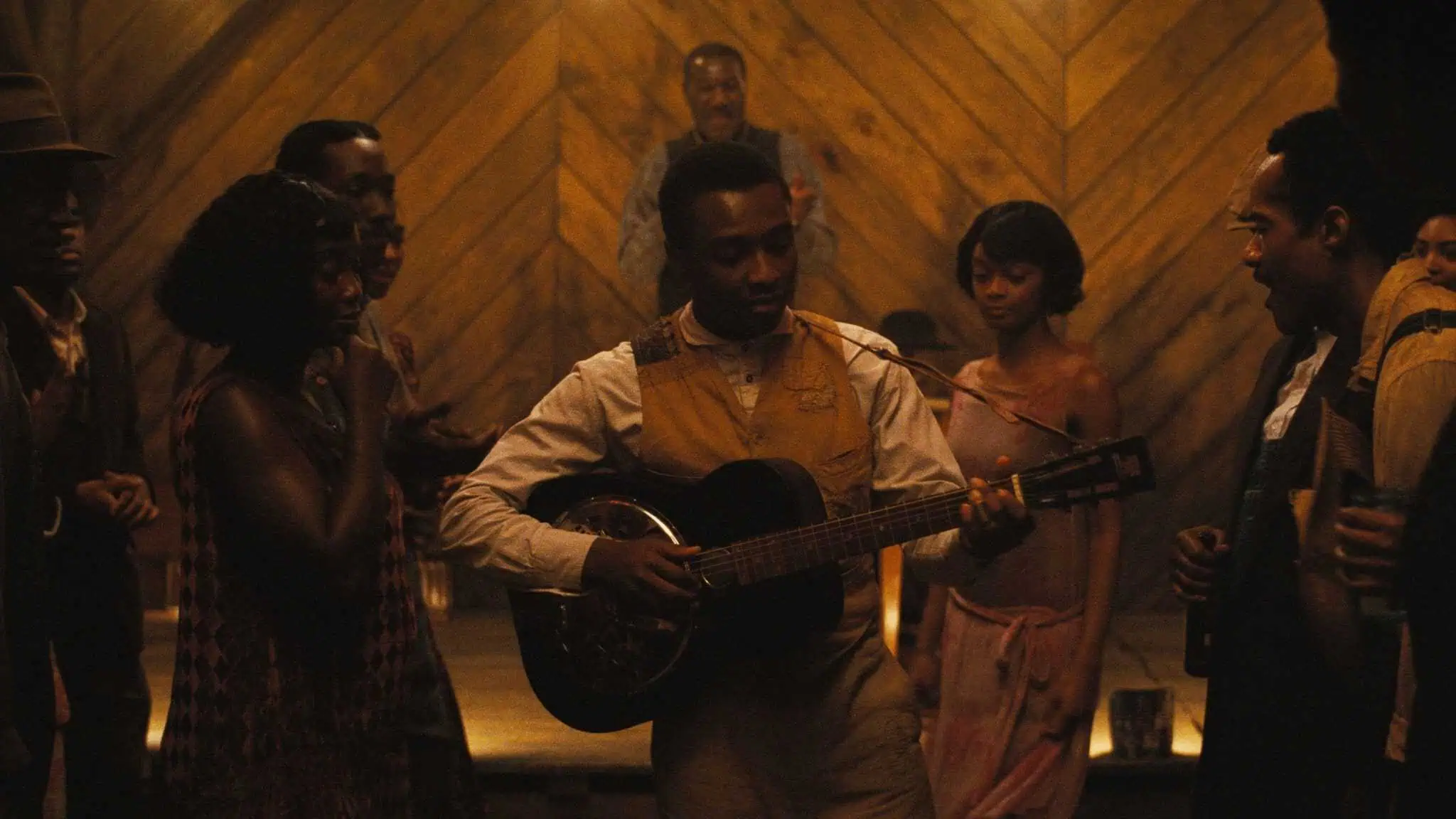 Still frame from "Sinners"; a man holds a guitar with people watching him play.