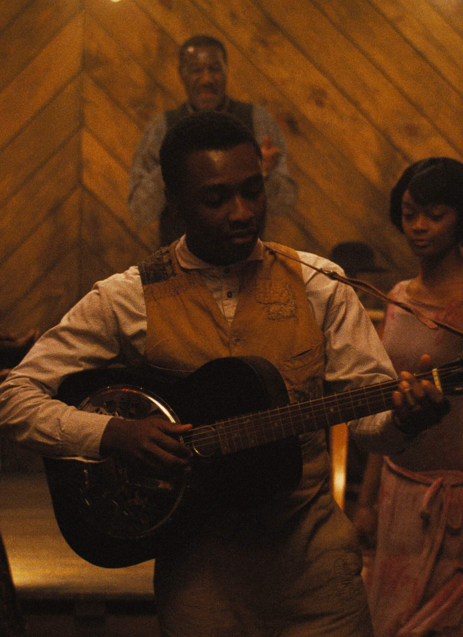 Still frame from "Sinners"; a man holds a guitar with people watching him play.