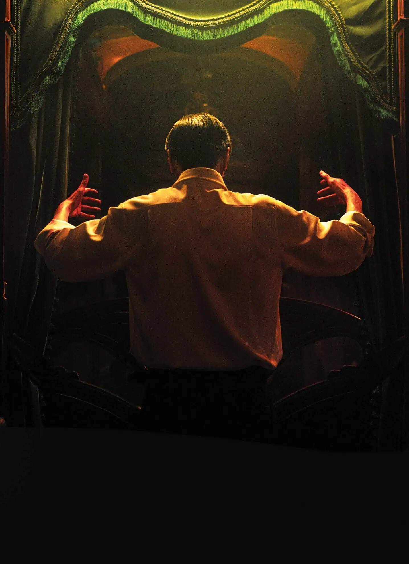 The back of a man with dark hair, in a white shirt, standing with his up, with a curtain and stage in the background.