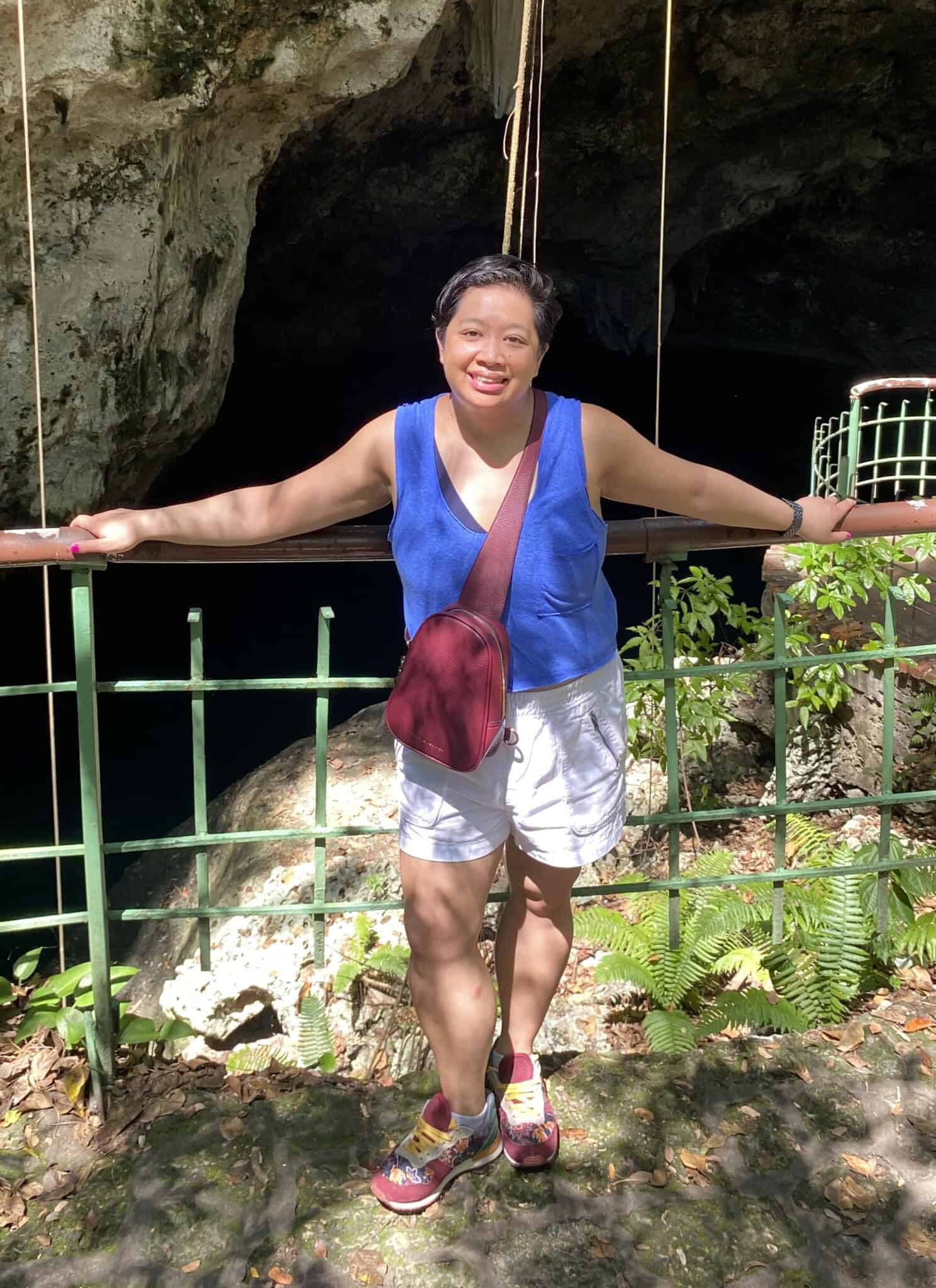 An Asian woman, in a blue top and white shorts and red bag, stands in front of dark cave.