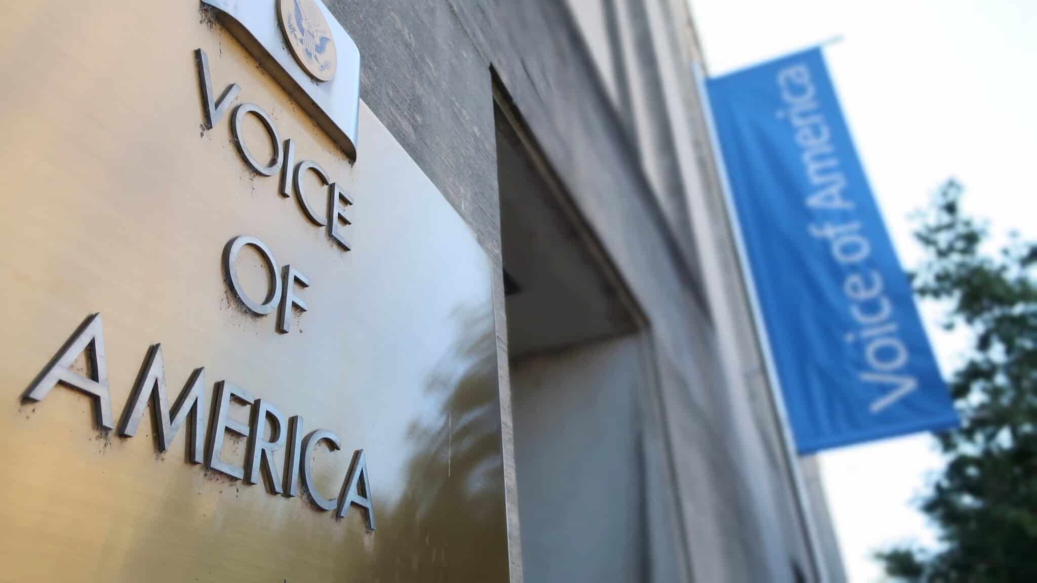 A dark gold sign that reads "Voice of America" on the side of a building, with a blue sign in the background.