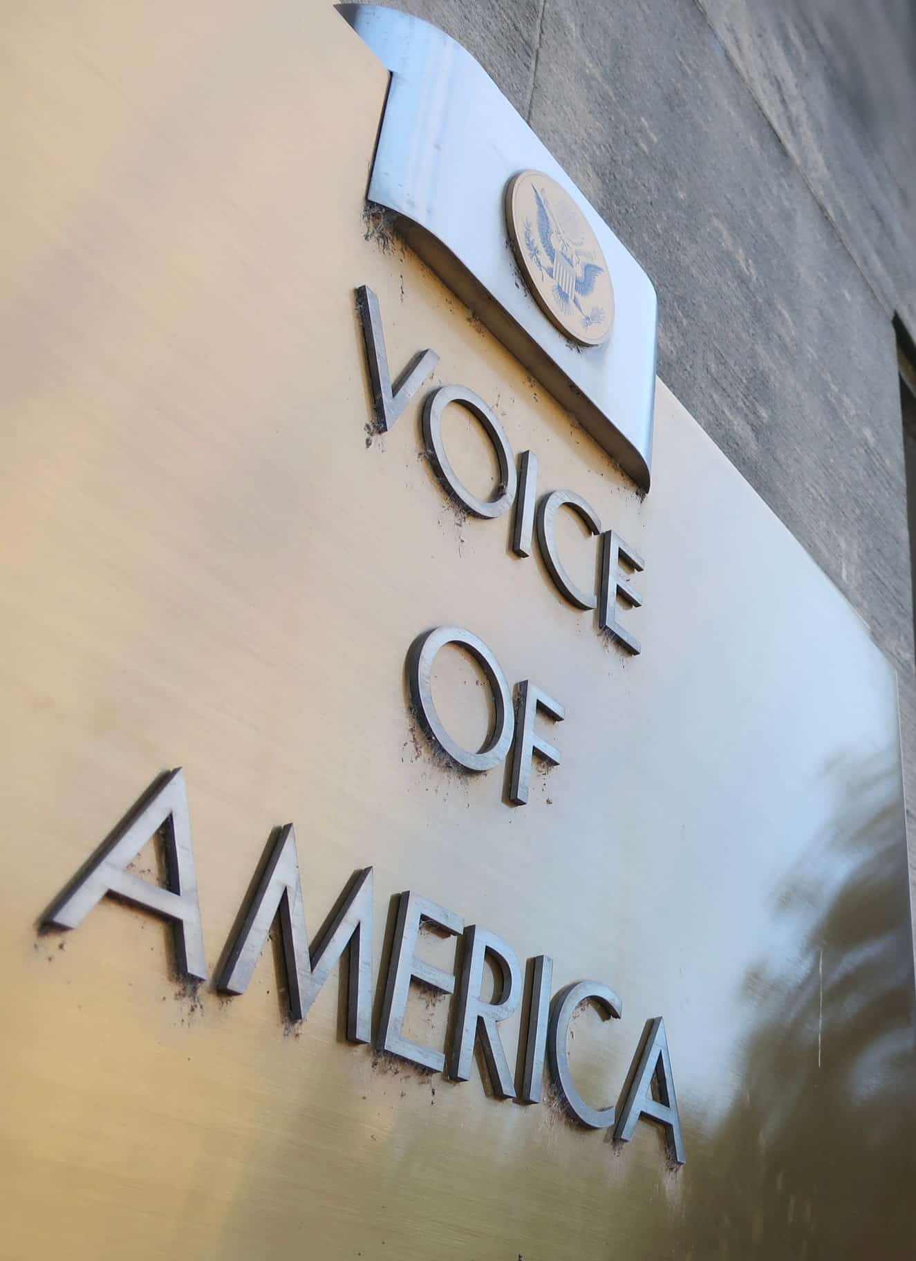 A dark gold sign that reads "Voice of America" on the side of a building, with a blue sign in the background.