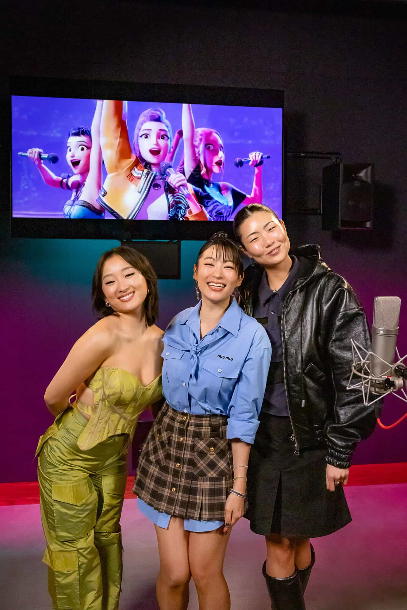 Three Asian women stand together smiling, one dressed in green, one in blue and one in black, with an animated film in the background.