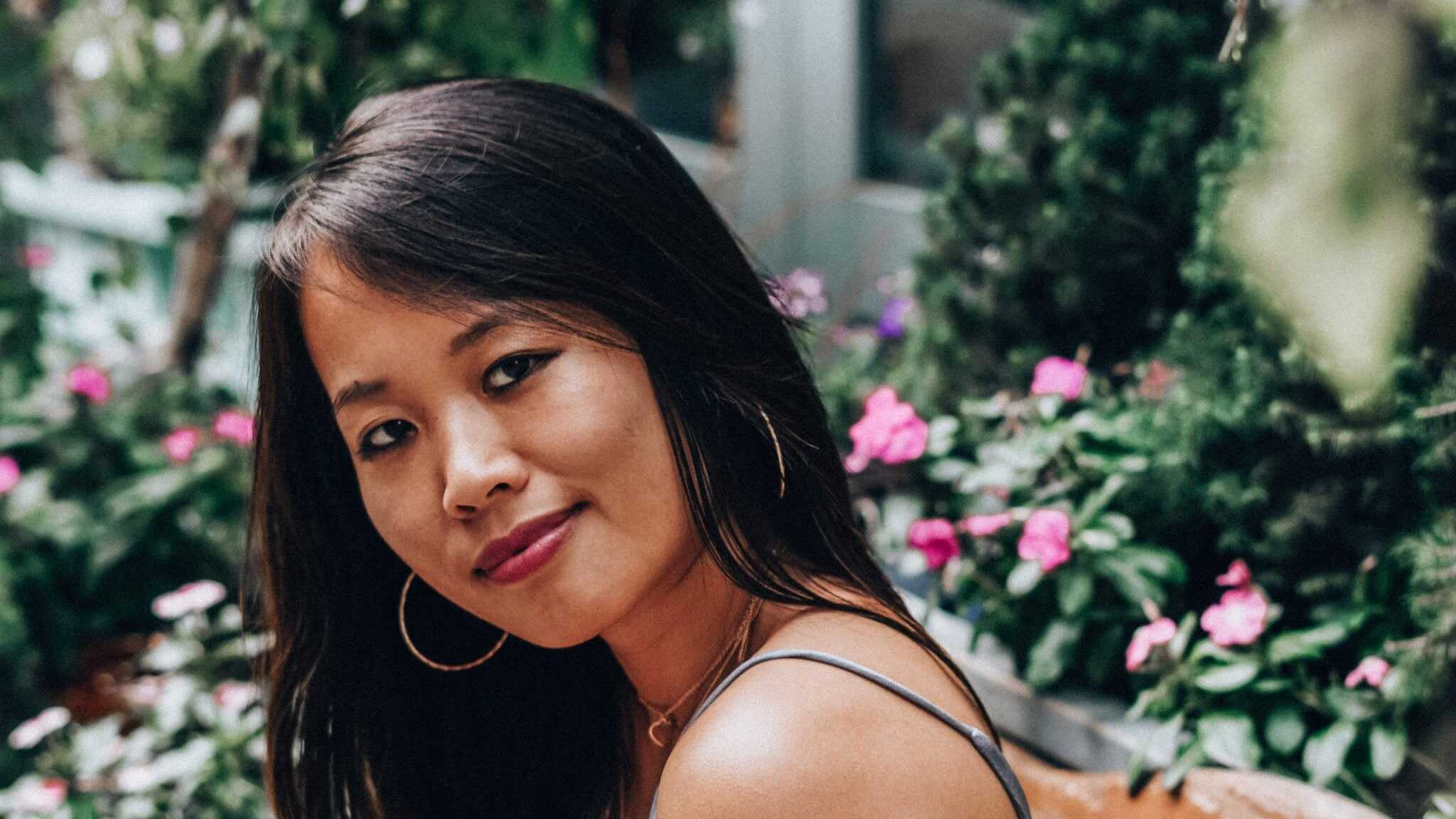 An Asian American woman, with long black hair, in a gray tank top, with flowers in the background.