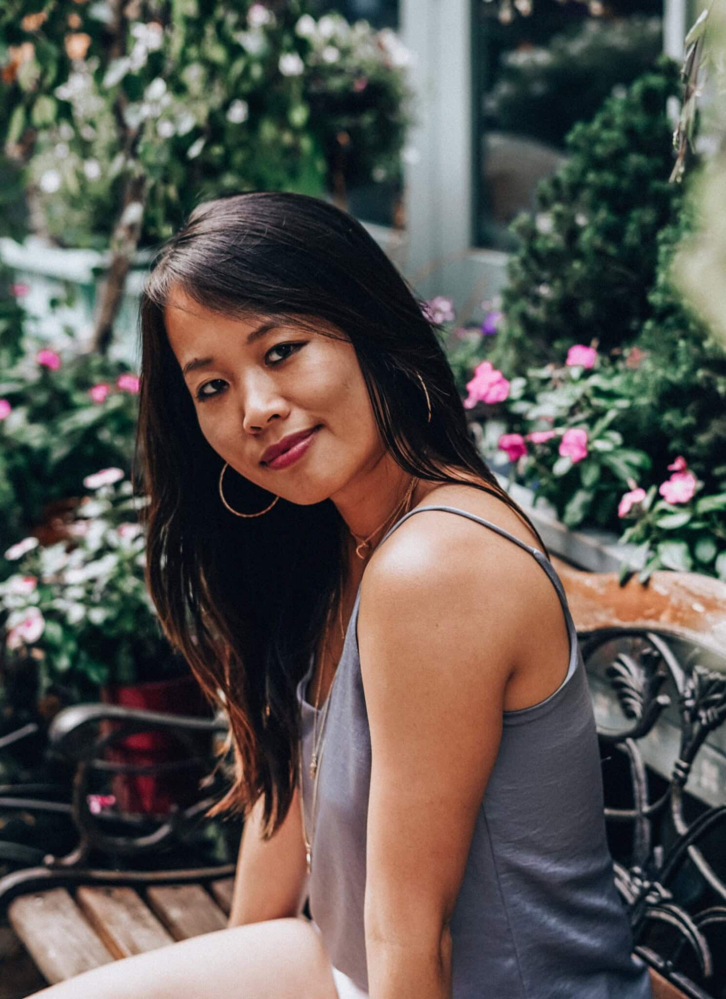 An Asian American woman, with long black hair, in a gray tank top, with flowers in the background.
