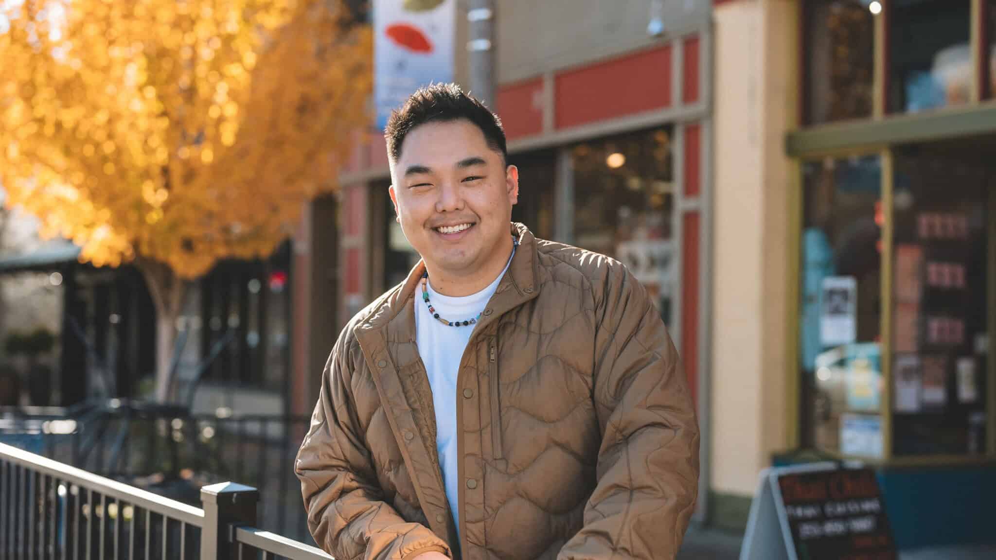 An Asian man in a brown jacket and white shirt stands next to black railing, with businesses in the background.