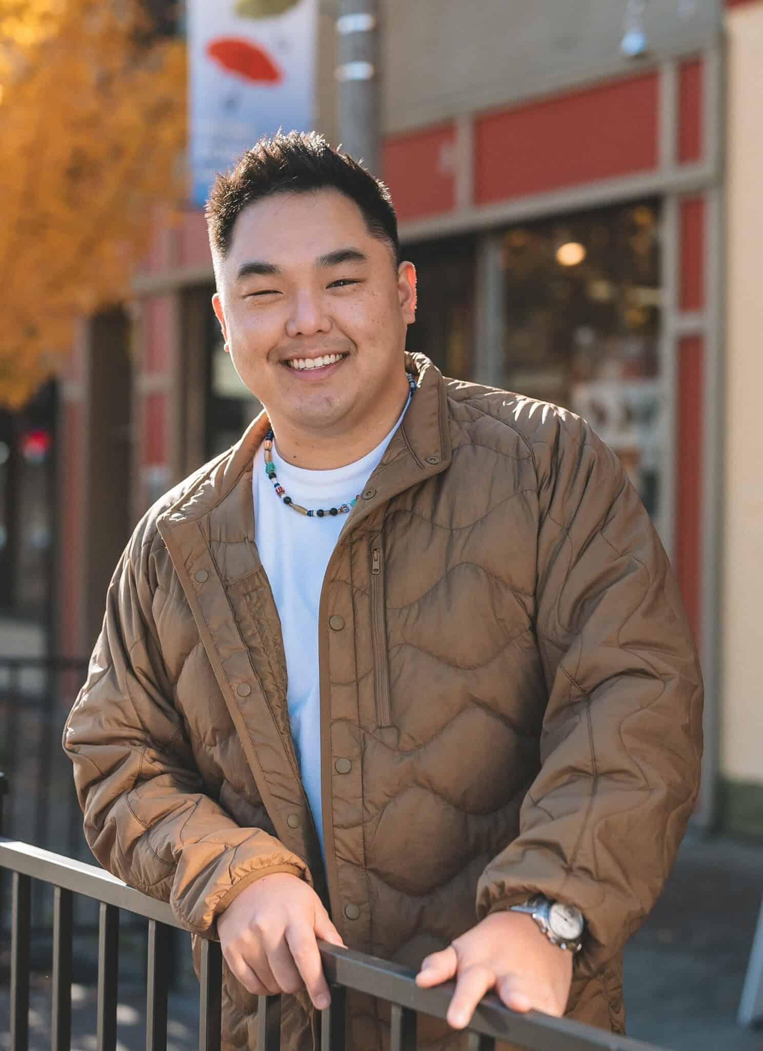 An Asian man in a brown jacket and white shirt stands next to black railing, with businesses in the background.