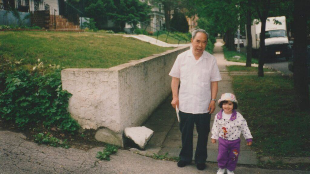 An older Asian man with a very young mixed-Asian girl stand together on a tree-lined sidewalk.