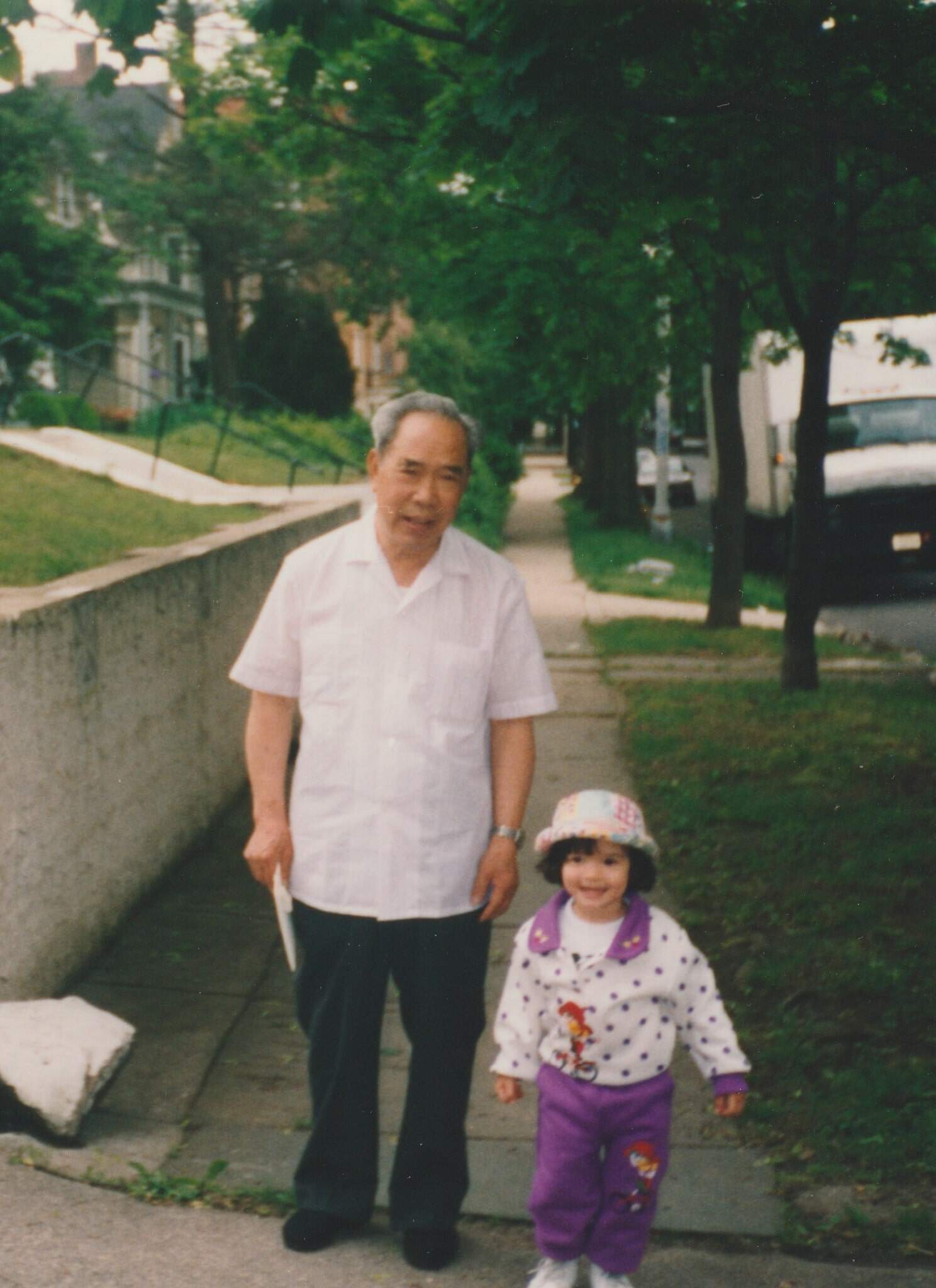 An older Asian man with a very young mixed-Asian girl stand together on a tree-lined sidewalk.