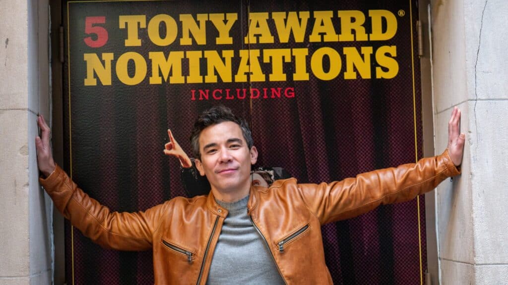 Conrad Ricamora stands below a sign that reads “5 Tony Award Nominations Including”
